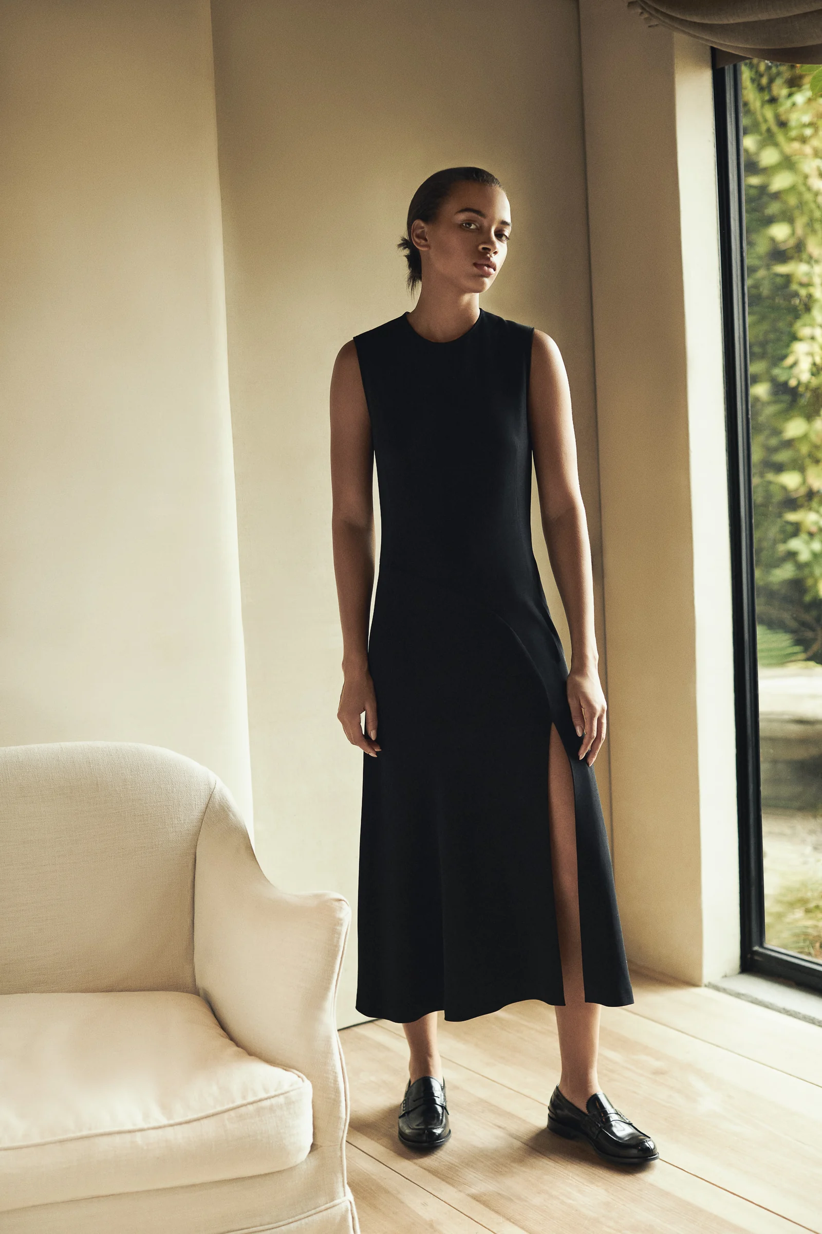 Model in a black sleeveless midi dress with side slit and loafers beside a cream armchair and window, for Another Tomorrow.