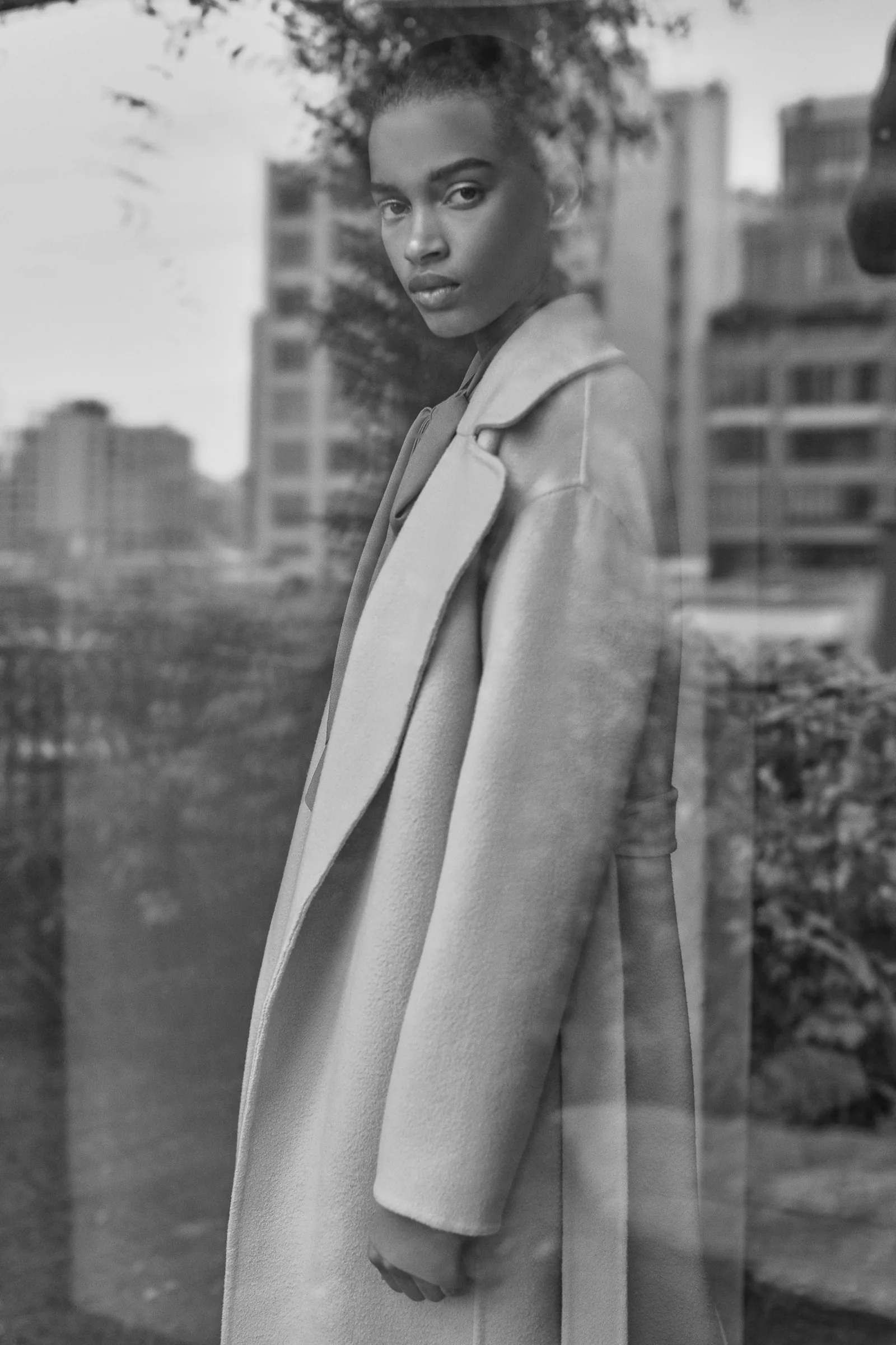 Black-and-white shot of a model in a long wool coat seen through a reflective window, city buildings behind, for Another Tomorrow.