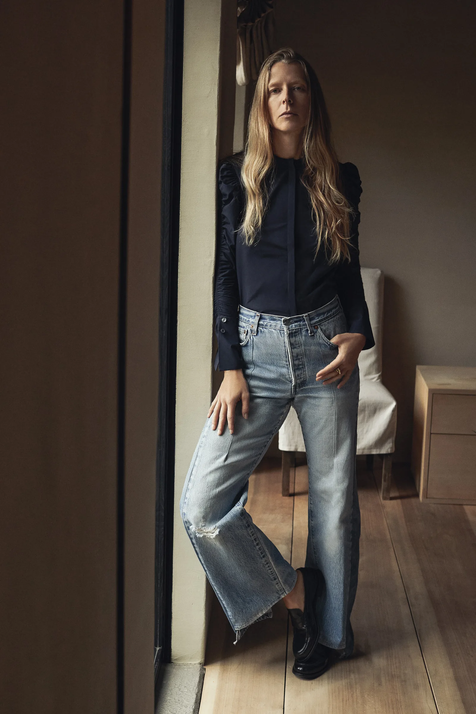 Woman in a navy puff-sleeve blouse and faded wide-leg jeans leaning by a window in a wood-floored room, for Another Tomorrow.