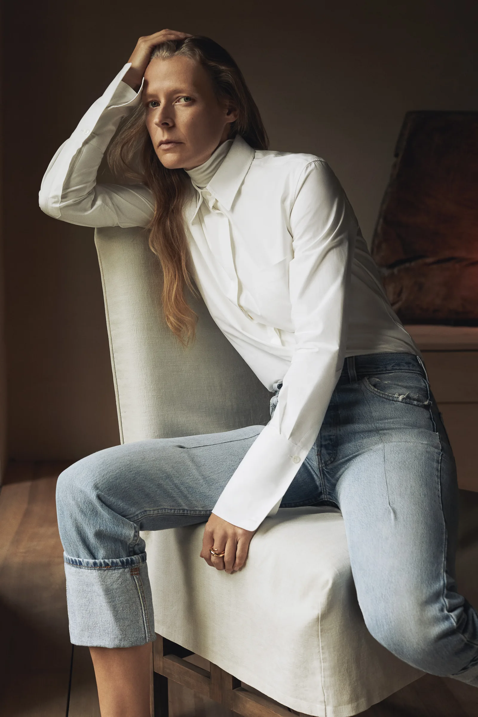 Woman in a white shirt, turtleneck and cuffed light-wash jeans perched on a linen chair, for Another Tomorrow.