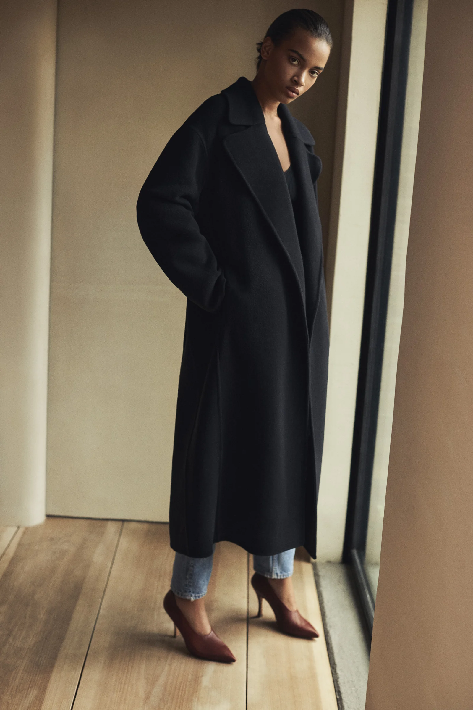Model in a long black wool coat, jeans and burgundy pointed heels standing by a window on wood floors, for Another Tomorrow.