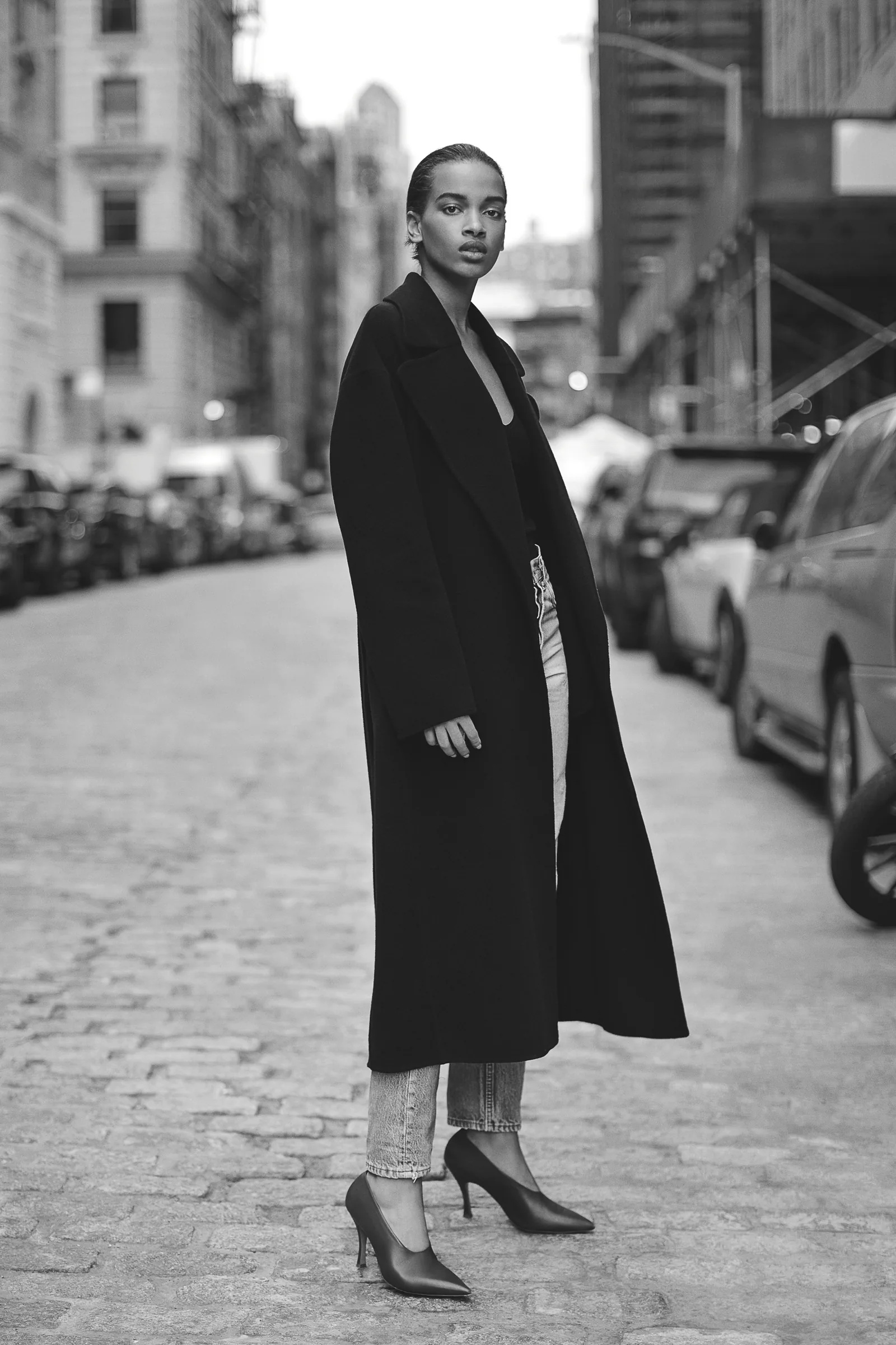 Black-and-white shot of a model in a long black coat, jeans and pointed heels on a cobblestone street, for Another Tomorrow.