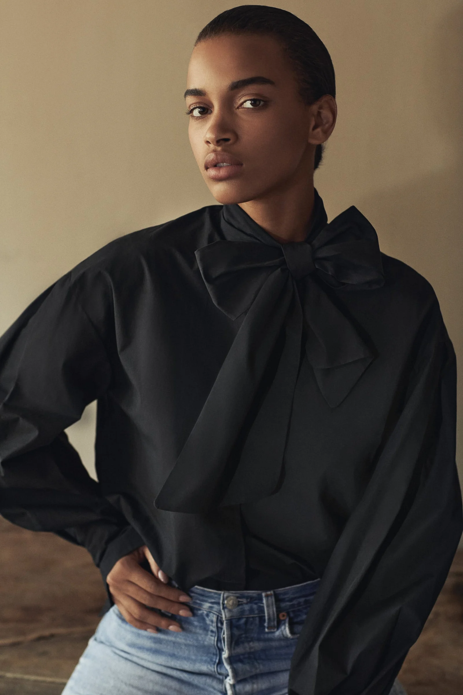 Model in a black pussy-bow blouse with voluminous sleeves and light blue jeans, hand on hip, for Another Tomorrow.