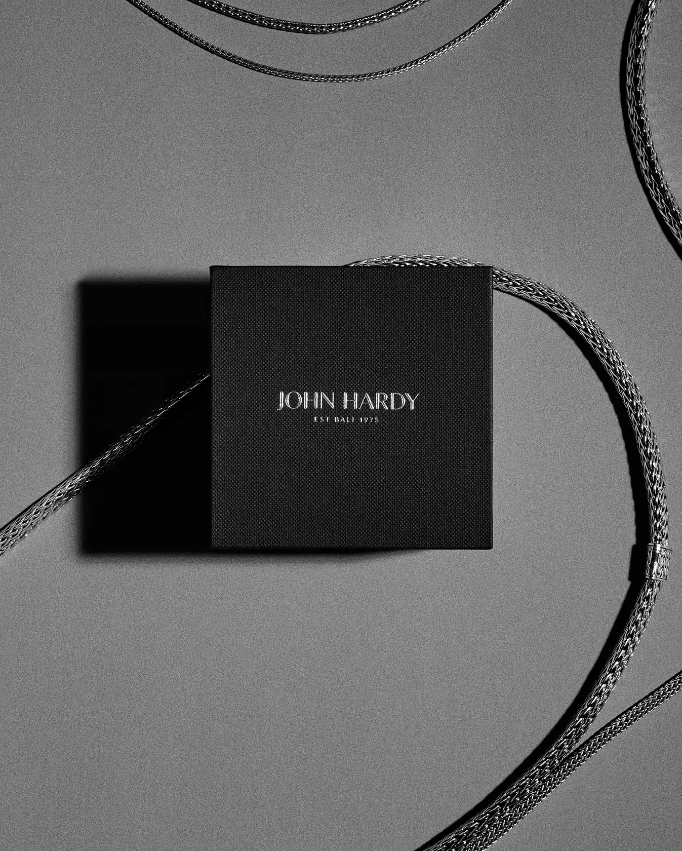 Black gift box with silver woven chain necklaces curving across a grey surface, in black and white, for John Hardy.