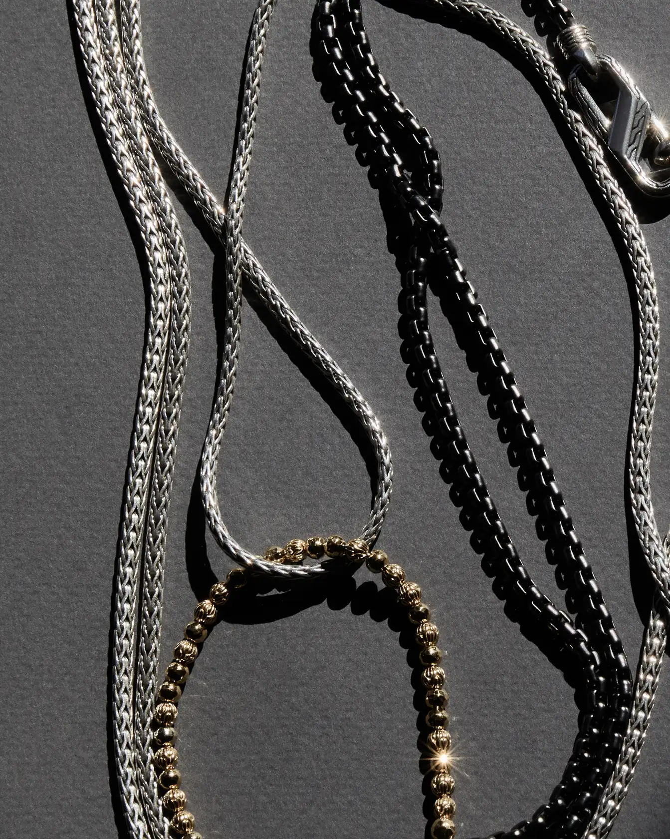 Silver woven chains, a black box chain and a gold bead bracelet on grey paper in hard sunlight, for John Hardy.