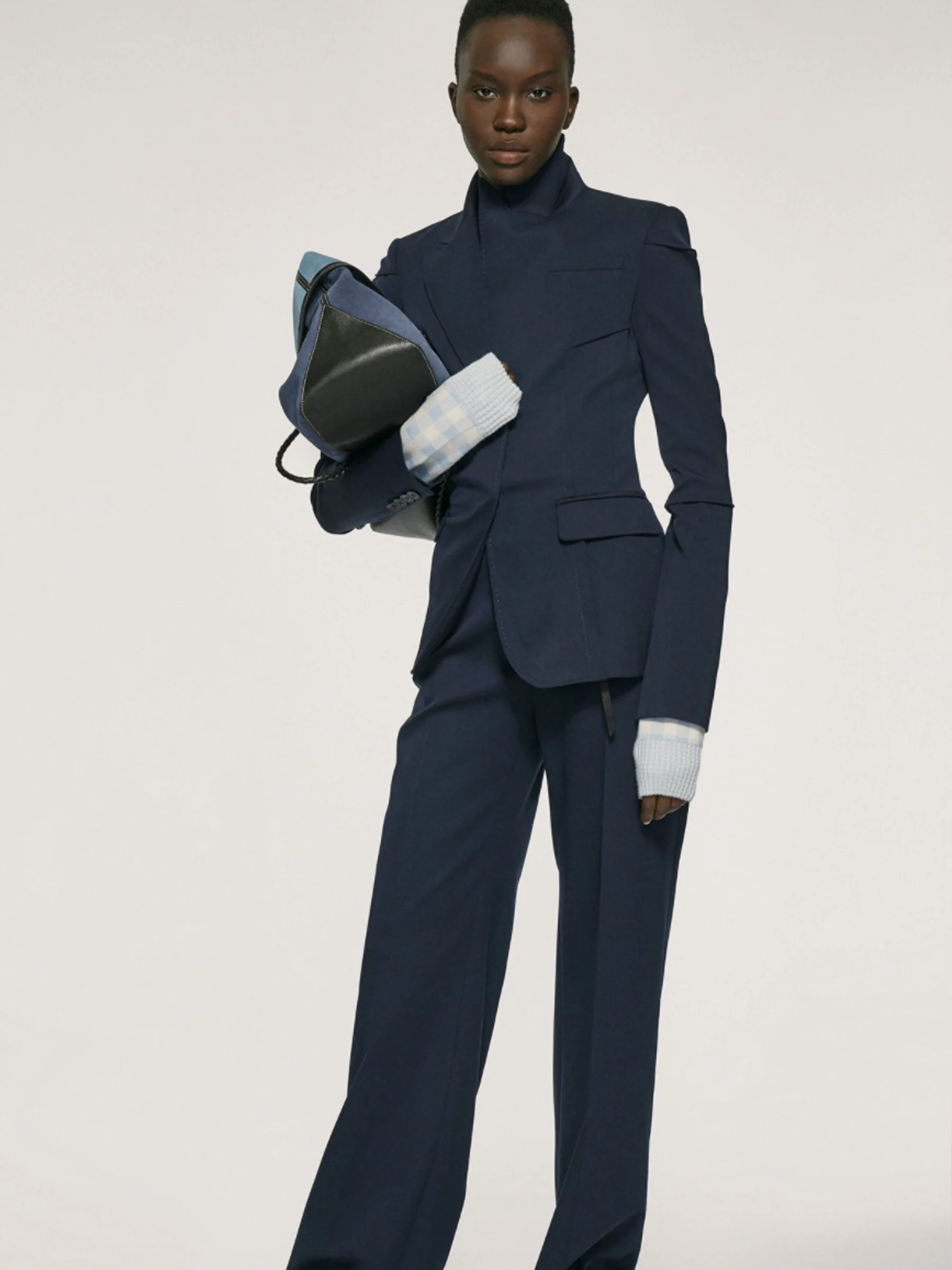 Model in a navy tailored suit with light blue cuffs, holding a denim and black leather bag, on a grey backdrop, for Altuzarra.