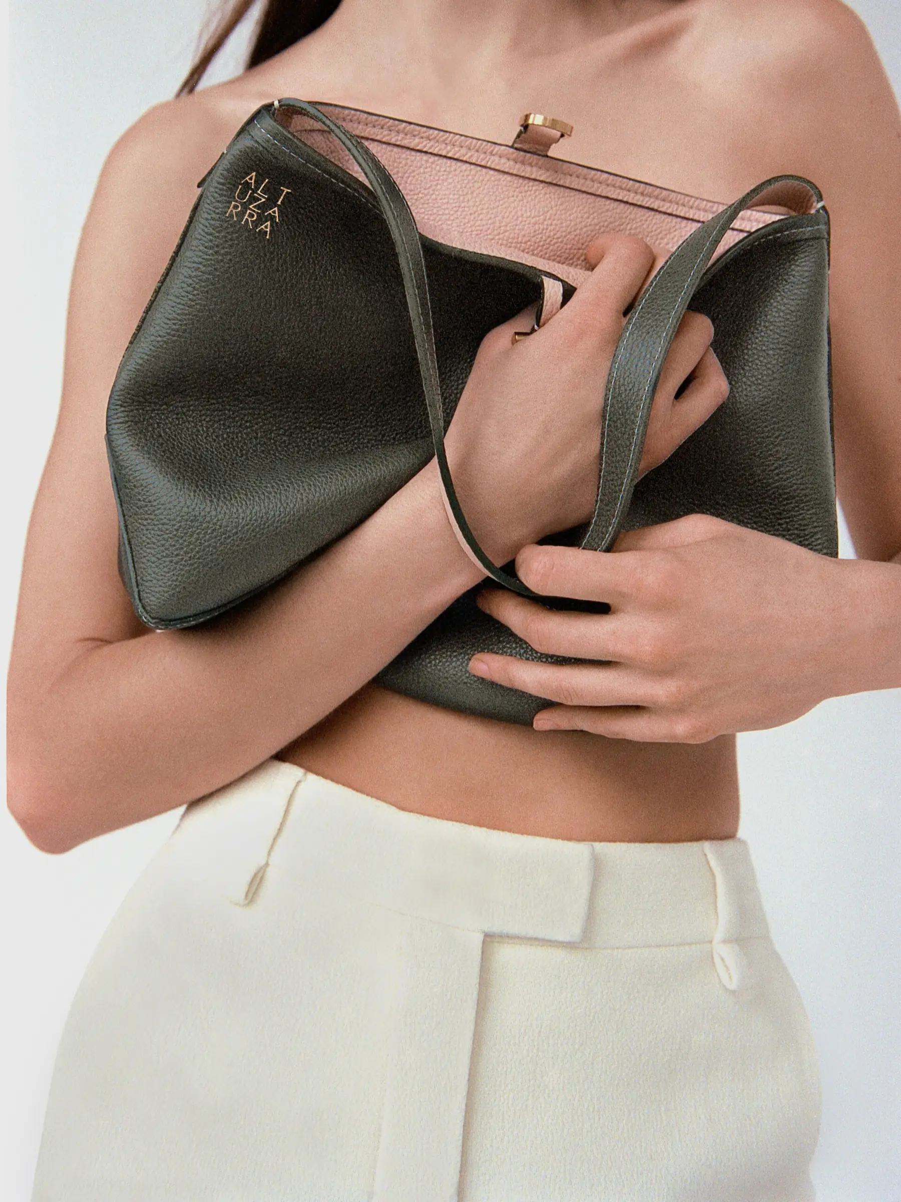 Model clutching an olive pebbled leather tote with pink lining against her bare torso, wearing cream trousers, for Altuzarra.