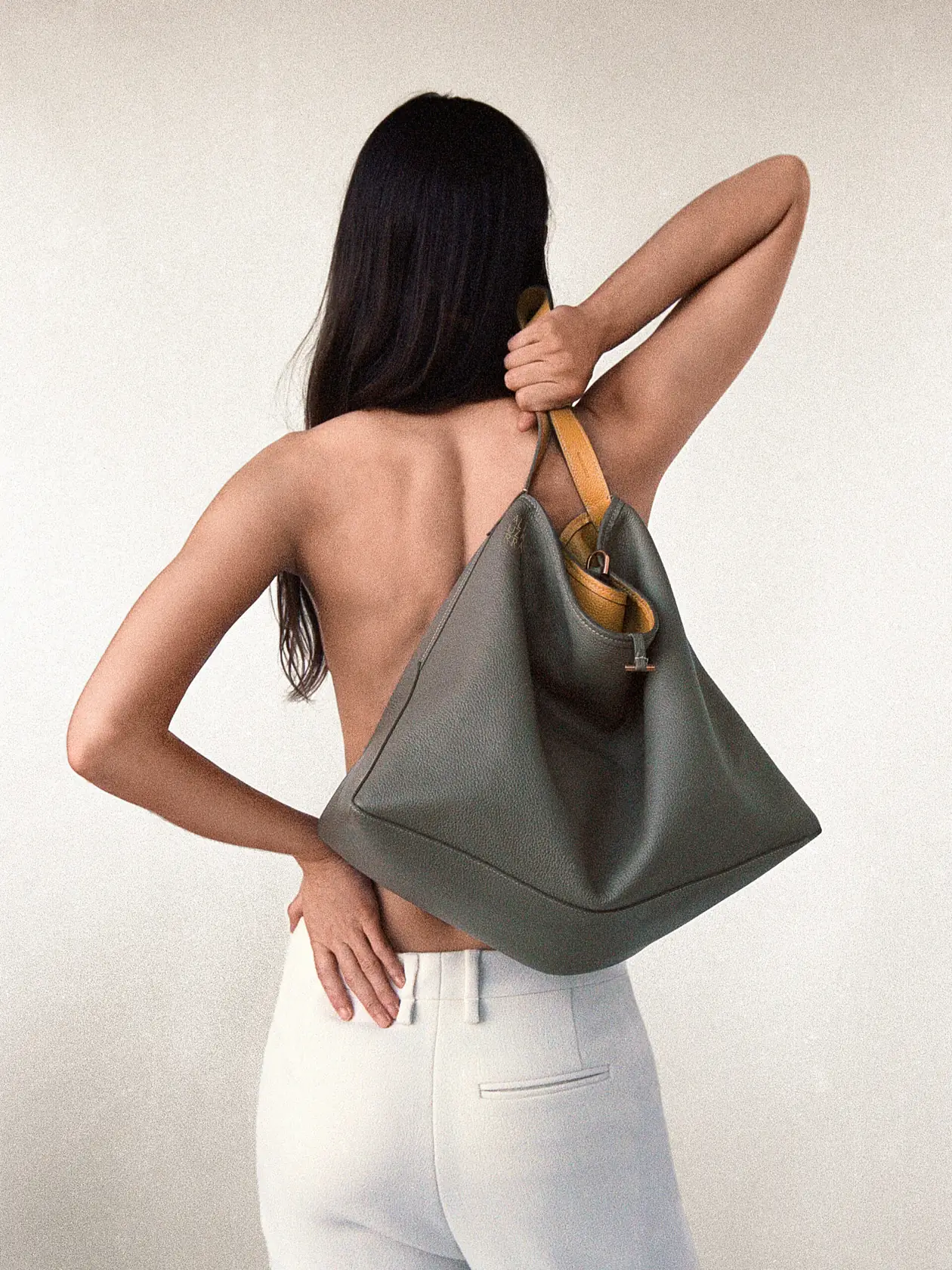 Model seen from behind with bare back, holding a grey leather bag with tan strap over her shoulder, white trousers, for Altuzarra.