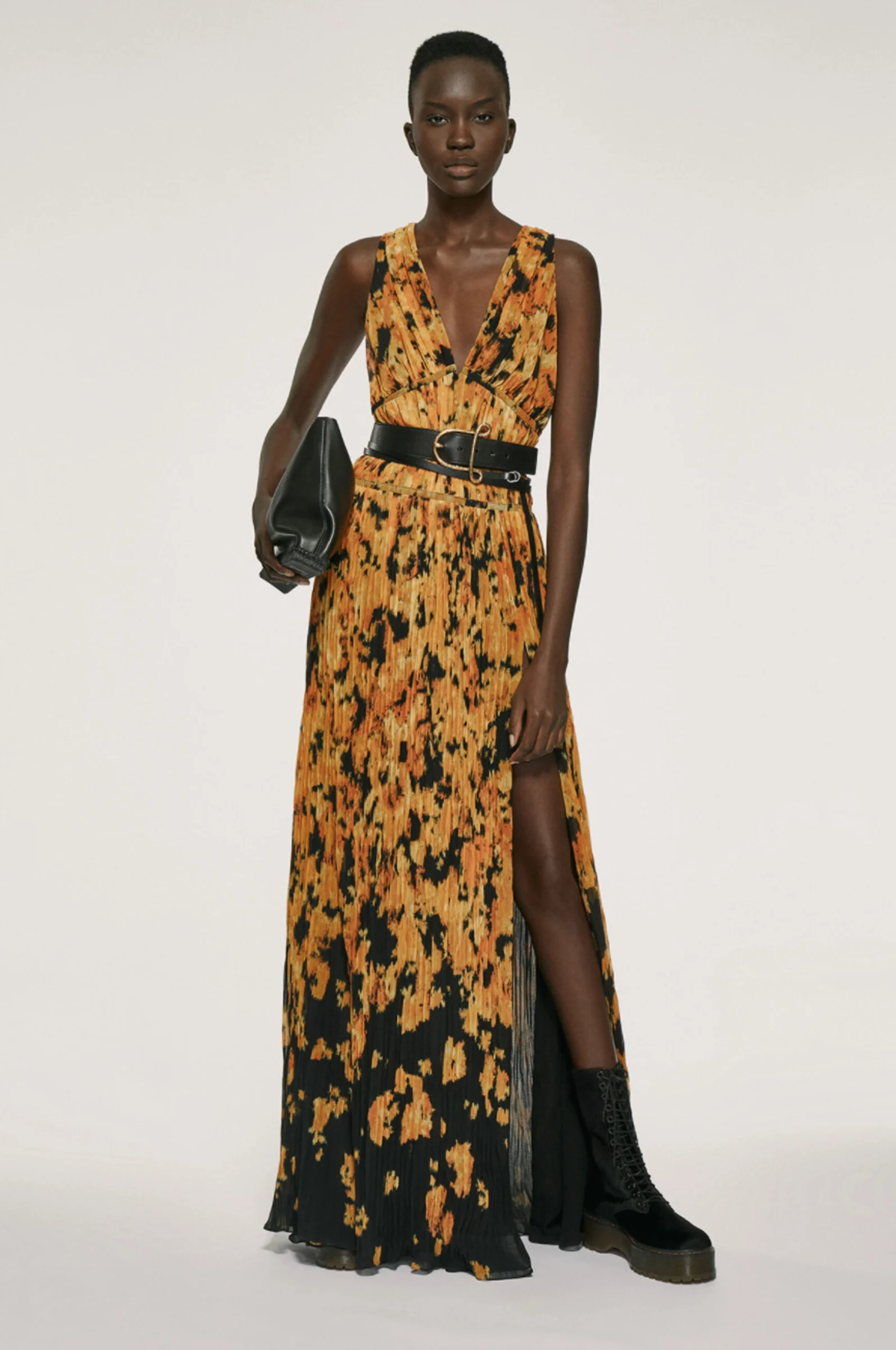 Model in a pleated orange and black printed maxi dress with black belt, clutch and lace-up boots, for Altuzarra.