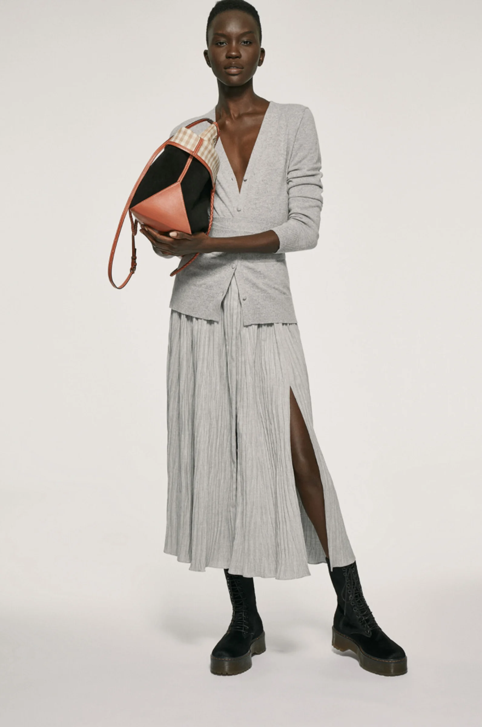 Model in grey cardigan and pleated slit skirt with black boots, holding a tan and black bucket bag, for Altuzarra.