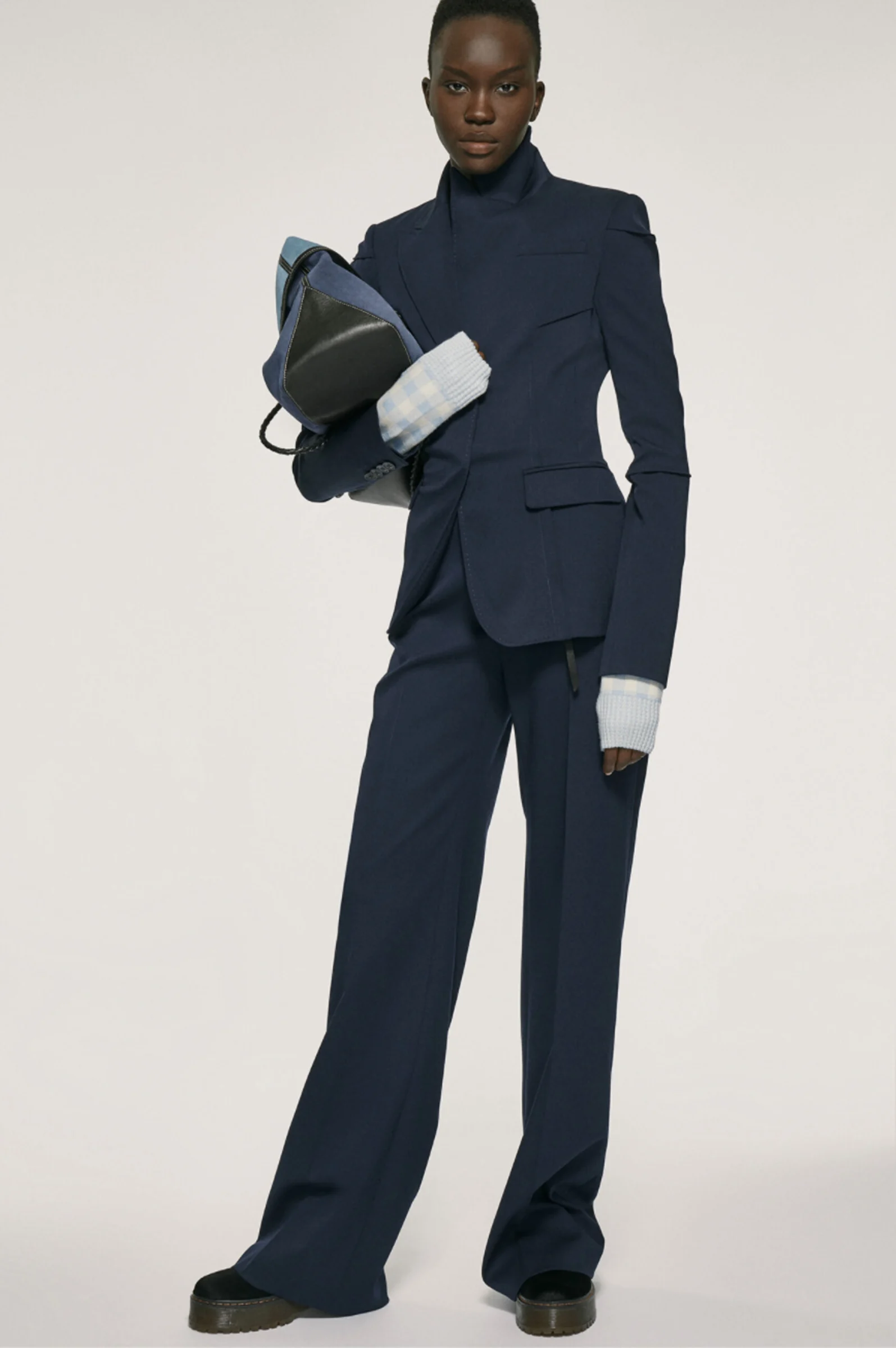 Model in a navy tailored blazer and wide-leg trousers holding a denim and leather bag on a pale backdrop, for Altuzarra.