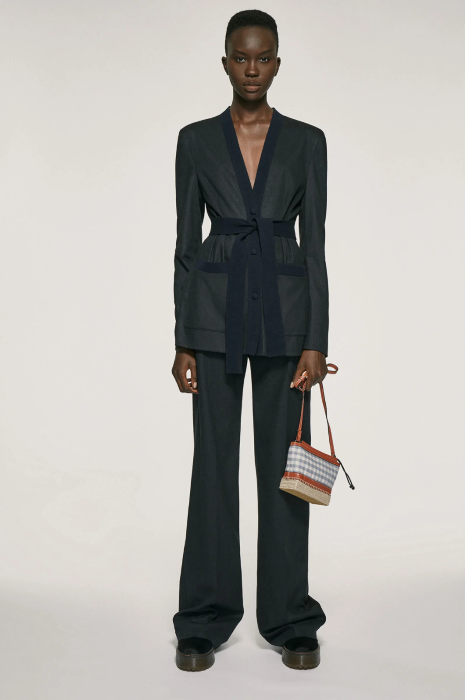 Model in a navy belted cardigan jacket and wide-leg trousers holding a gingham mini bag on grey backdrop, for Altuzarra.