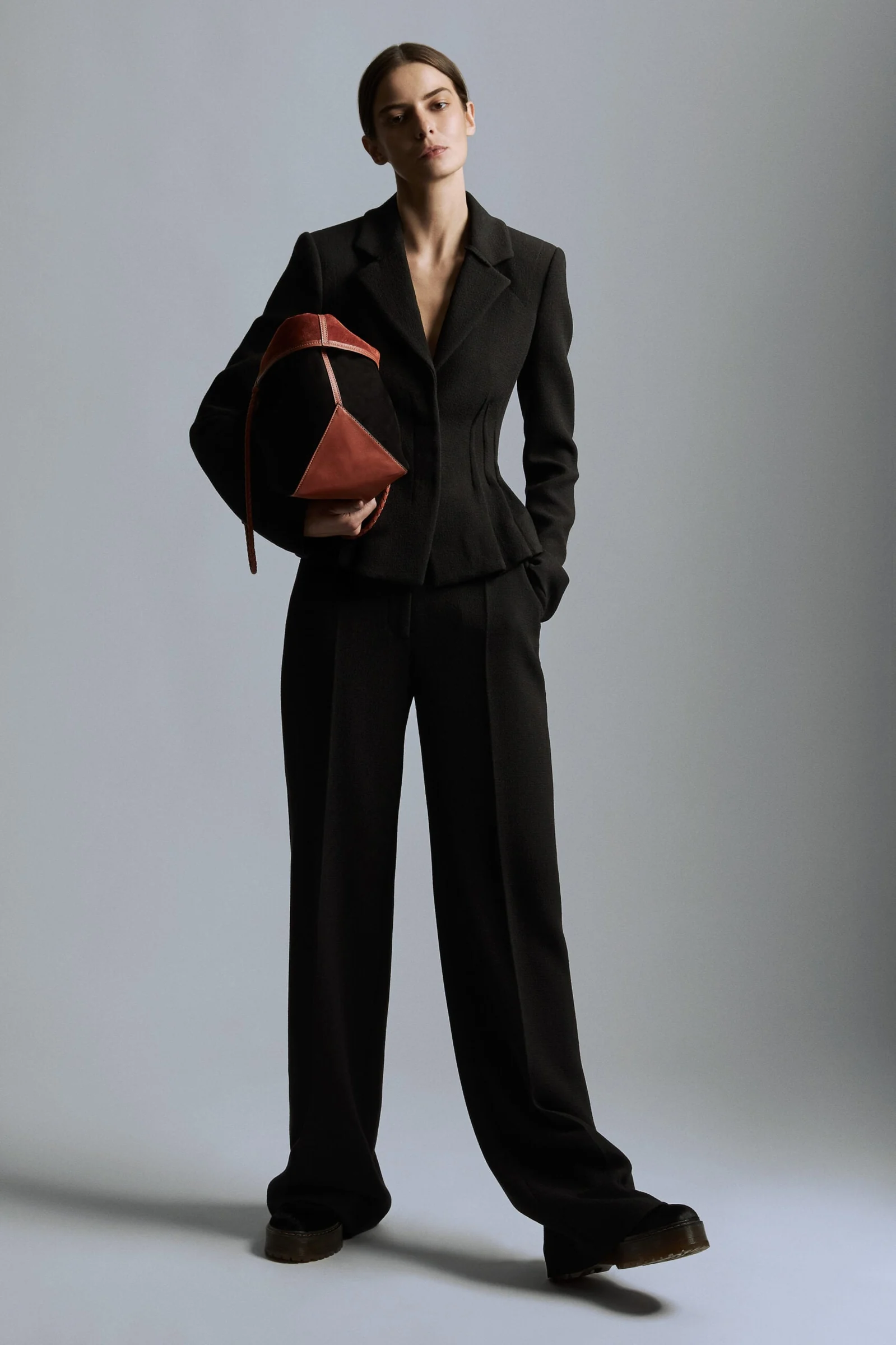 Model in a fitted black blazer and wide-leg trousers holding a red leather bag against a grey studio backdrop, for Altuzarra.