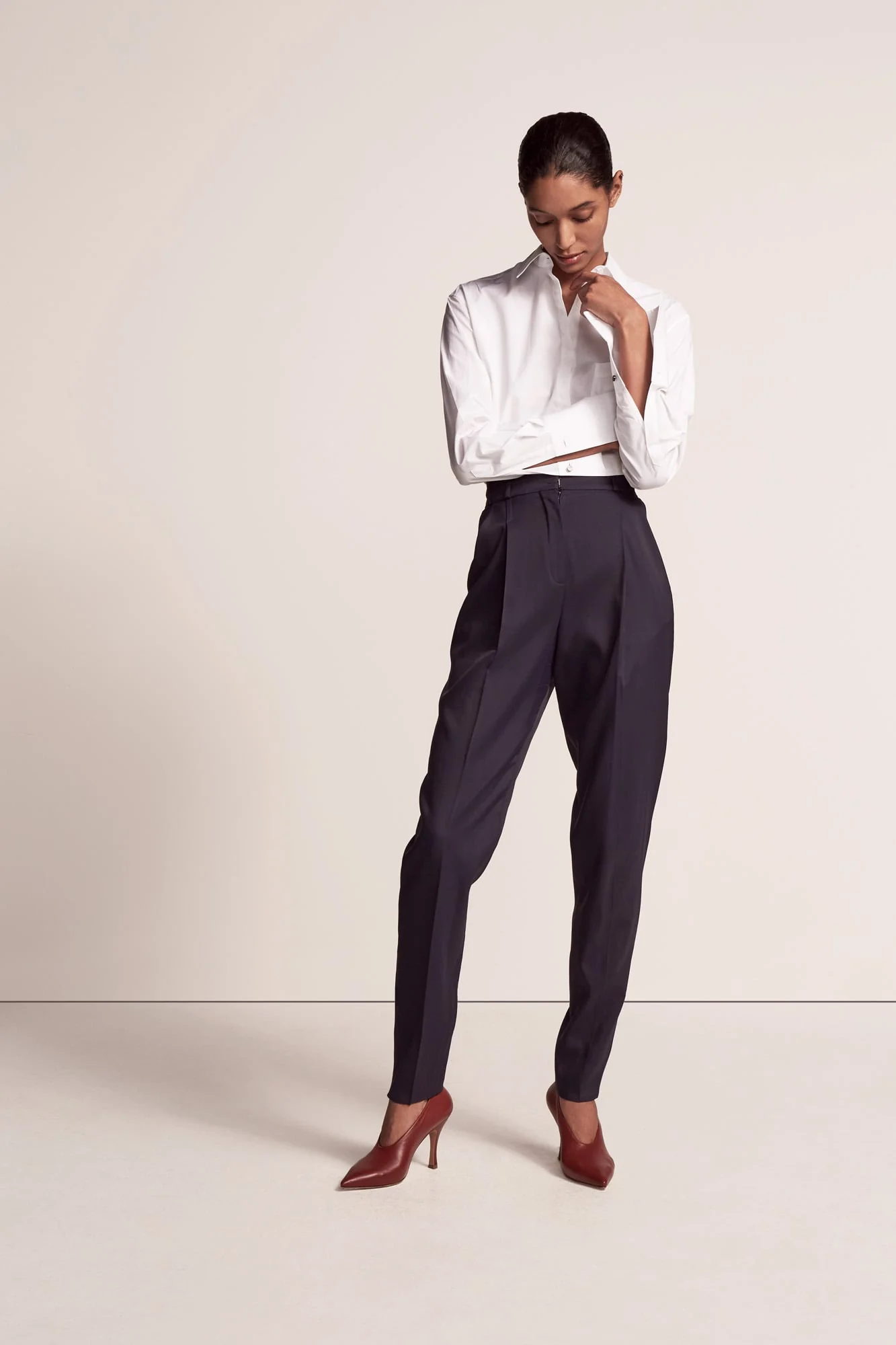 Model in a cropped white shirt, high-waisted navy pleated trousers and burgundy heels on a cream backdrop, for Another Tomorrow.