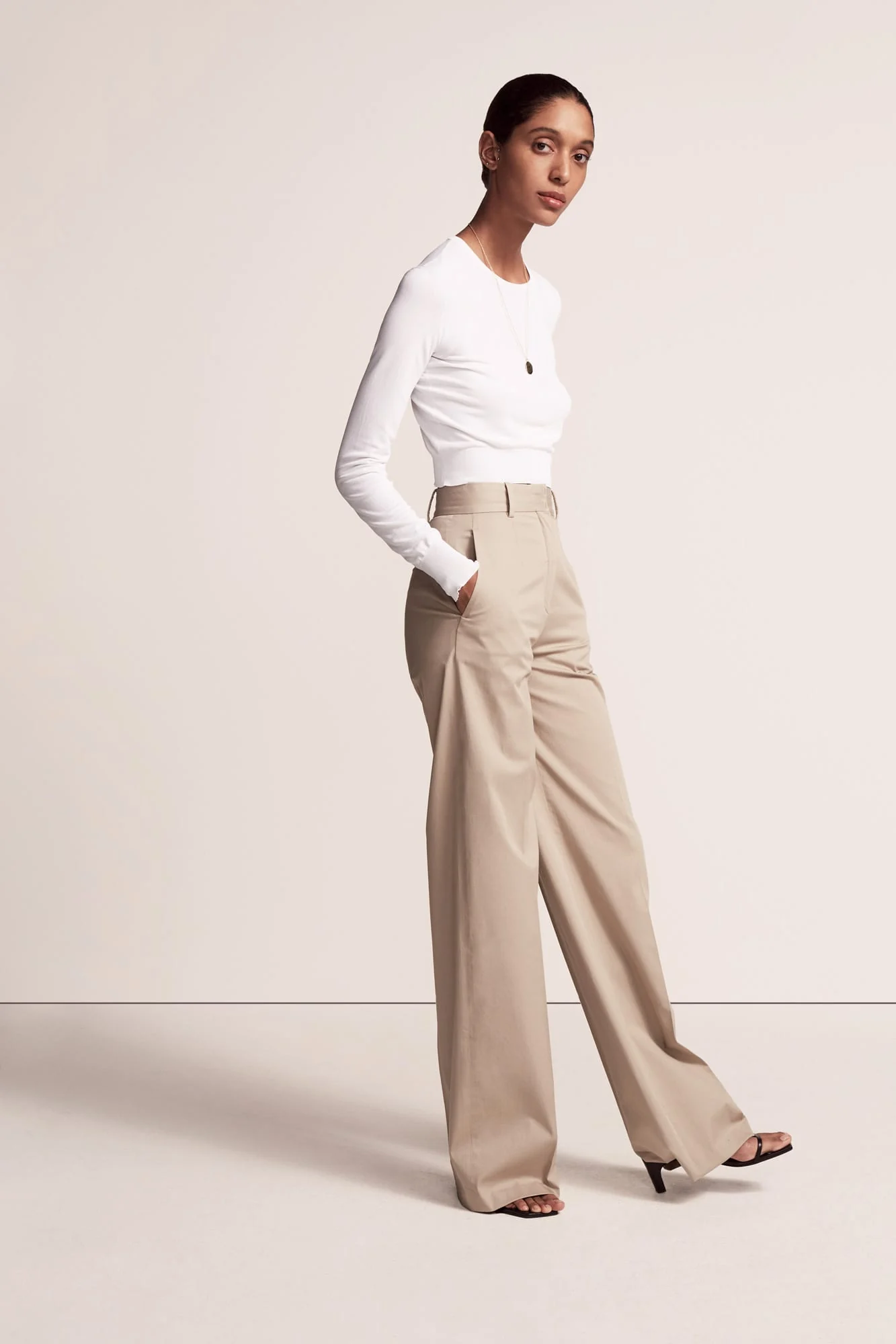Model in a white long-sleeve top and wide-leg khaki trousers on a plain cream studio set, for Another Tomorrow.