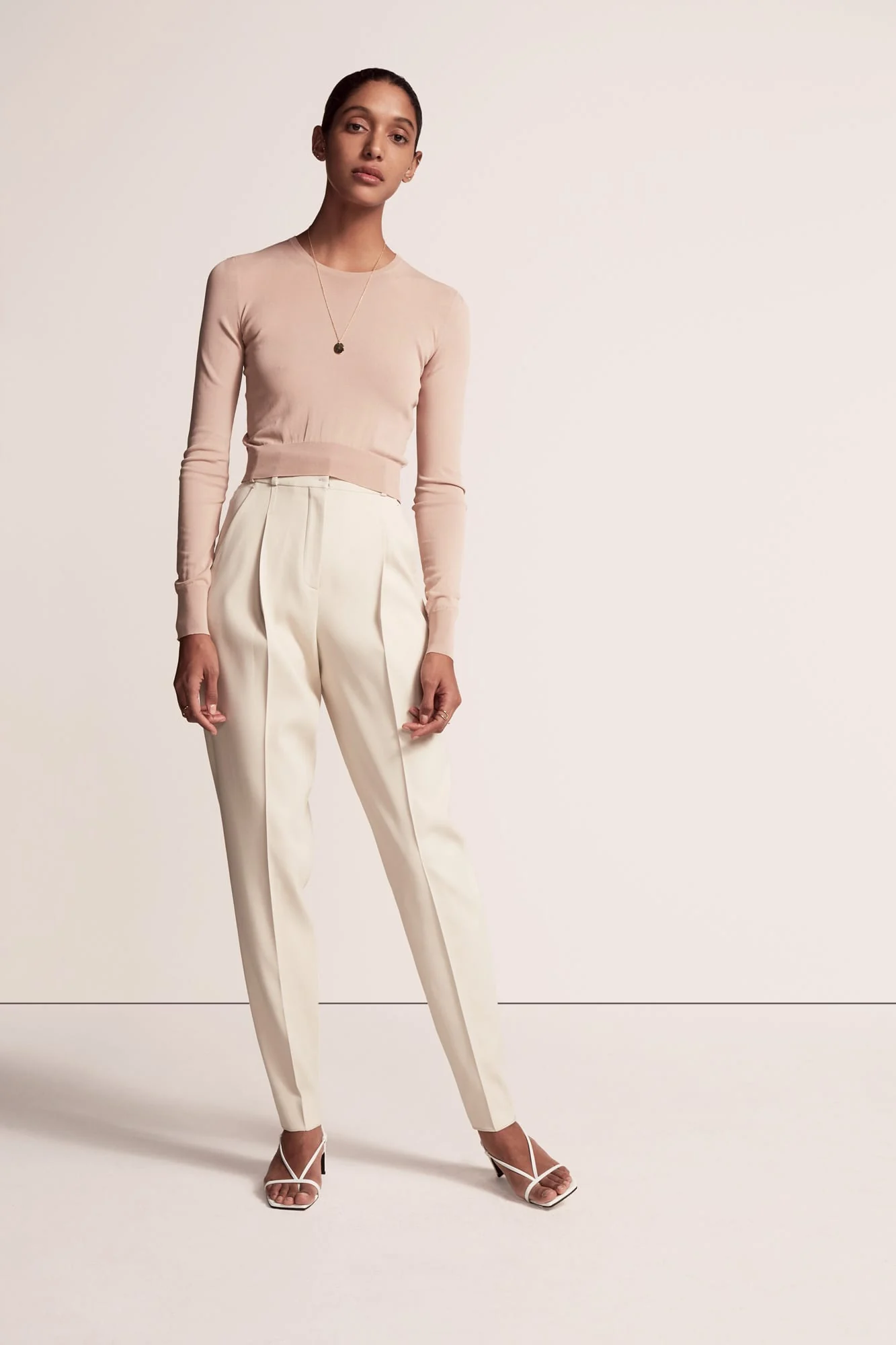 Model in a blush knit top, cream pleated trousers and white strappy sandals on a pale studio backdrop, for Another Tomorrow.