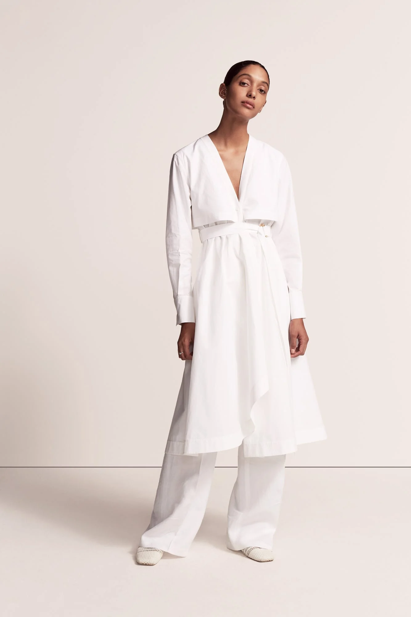 Model in a white belted cotton shirt dress over wide-leg white trousers on a plain studio backdrop, for Another Tomorrow.