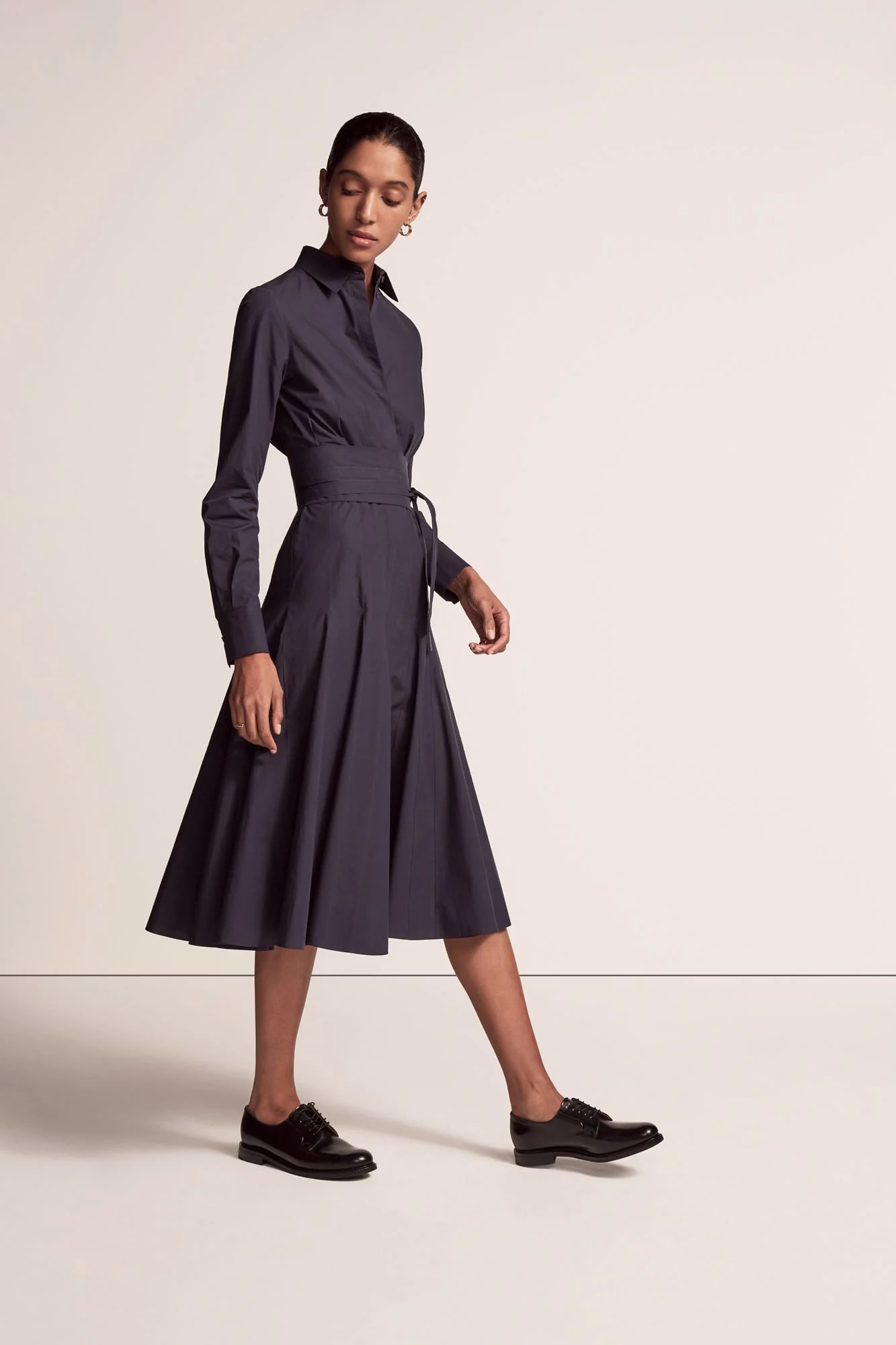 Model in a navy belted shirt dress with a full midi skirt and black leather oxfords on a light backdrop, for Another Tomorrow.