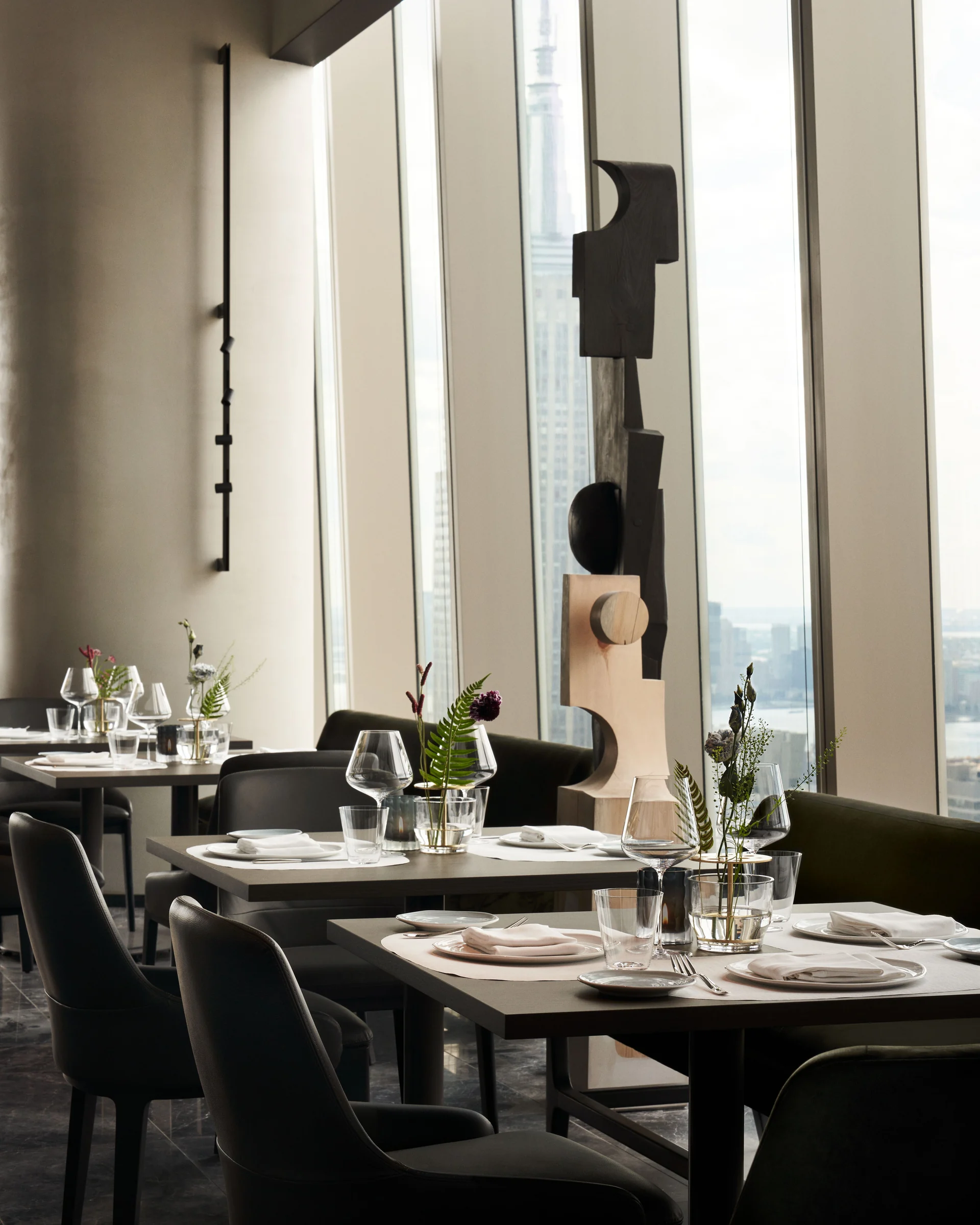 Restaurant dining room with set tables, wooden sculpture and tall windows facing the Manhattan skyline, for Centurion New York.
