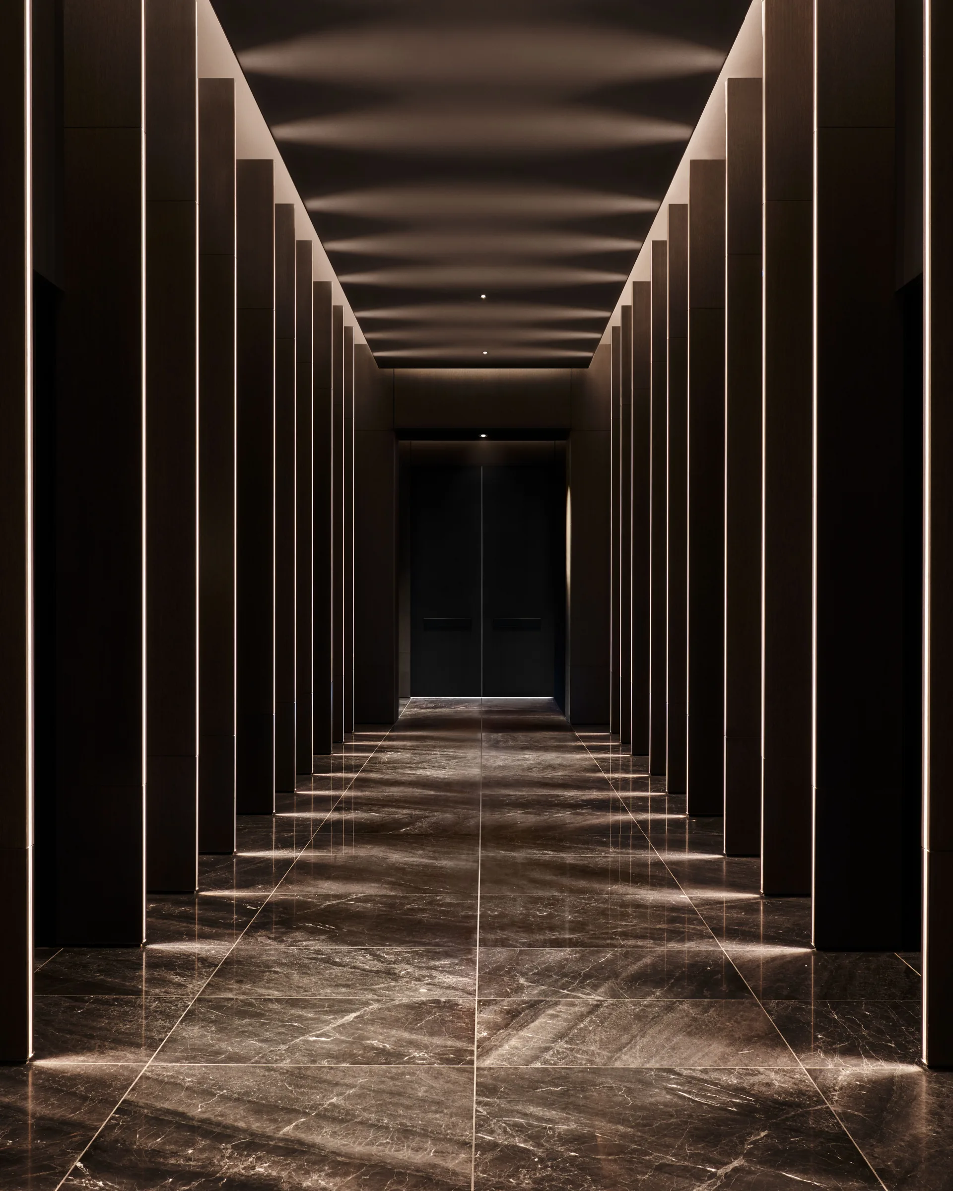 Corridor with polished dark marble floor, wood-panelled pillars and linear lighting leading to elevator doors, for Centurion New York.