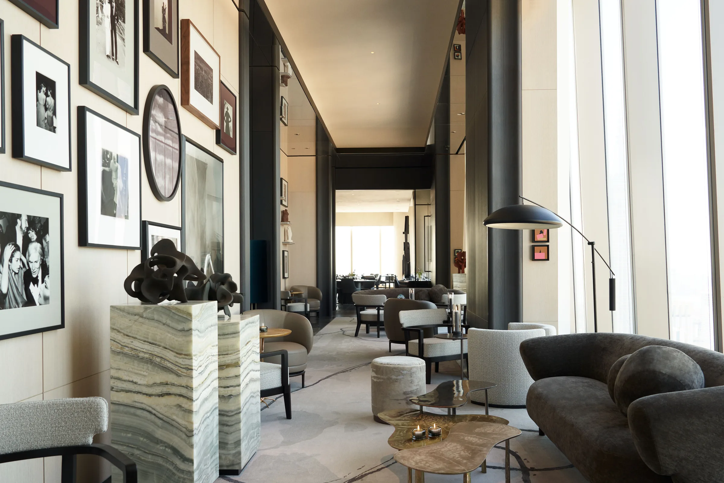 Lounge with framed photo gallery wall, onyx plinths, grey sofa and brass tables beside tall windows, for Centurion New York.