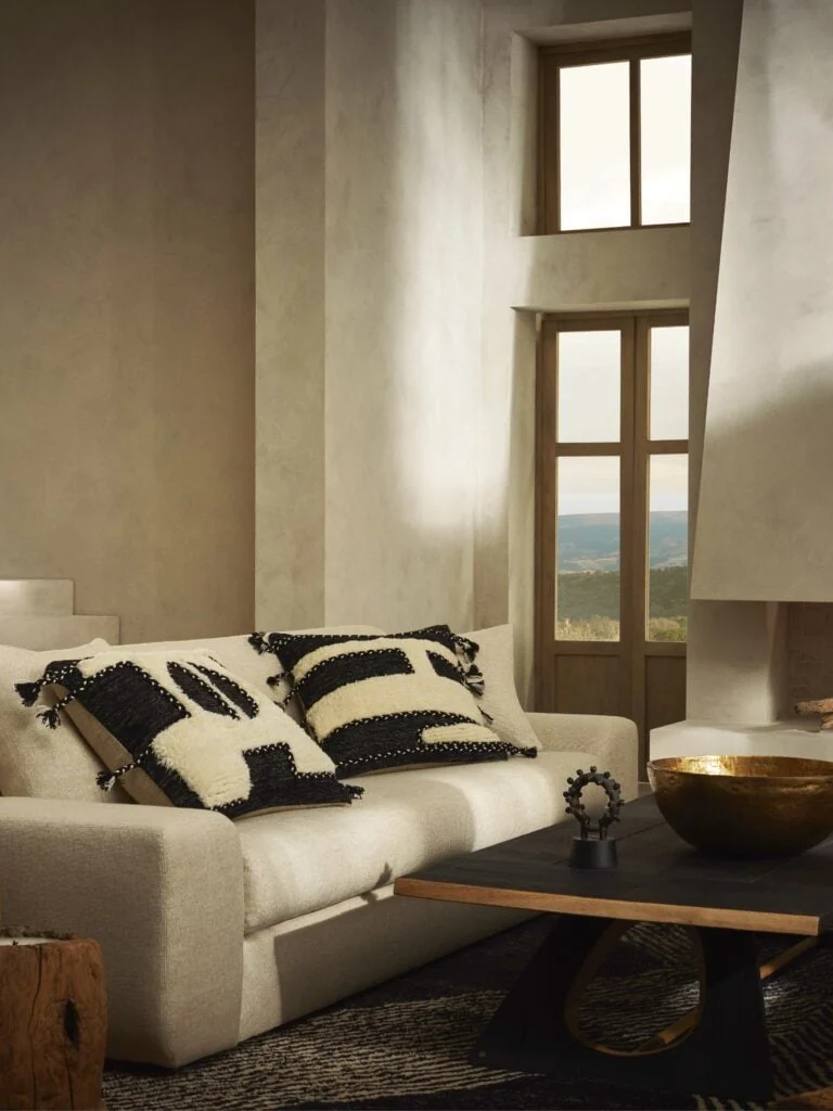 Linen sofa with black and cream wool pillows beside a dark coffee table and brass bowl in a plaster room, for Banana Republic.