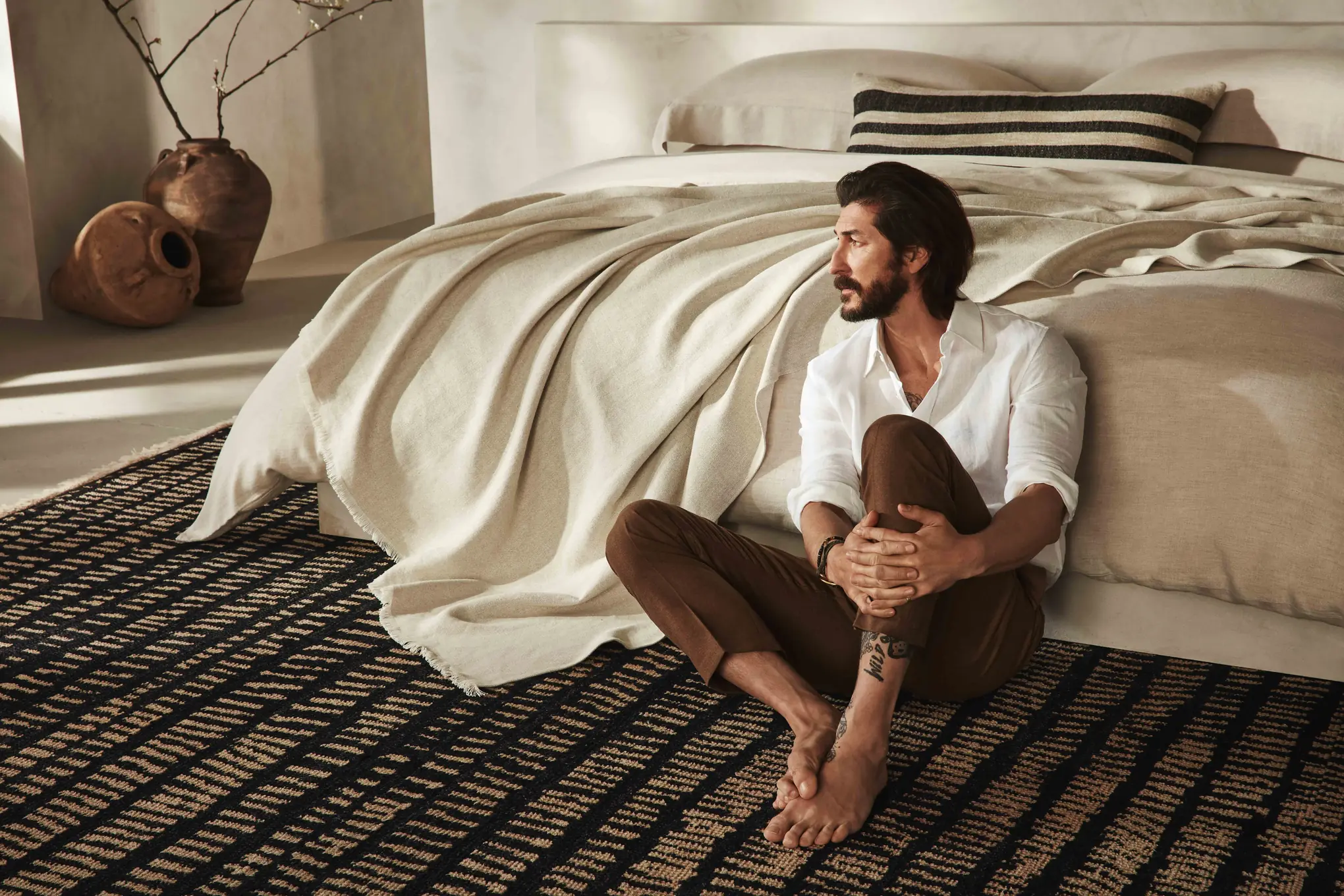 Barefoot man in white shirt sits on a woven rug beside a bed draped in a linen throw, for Banana Republic.
