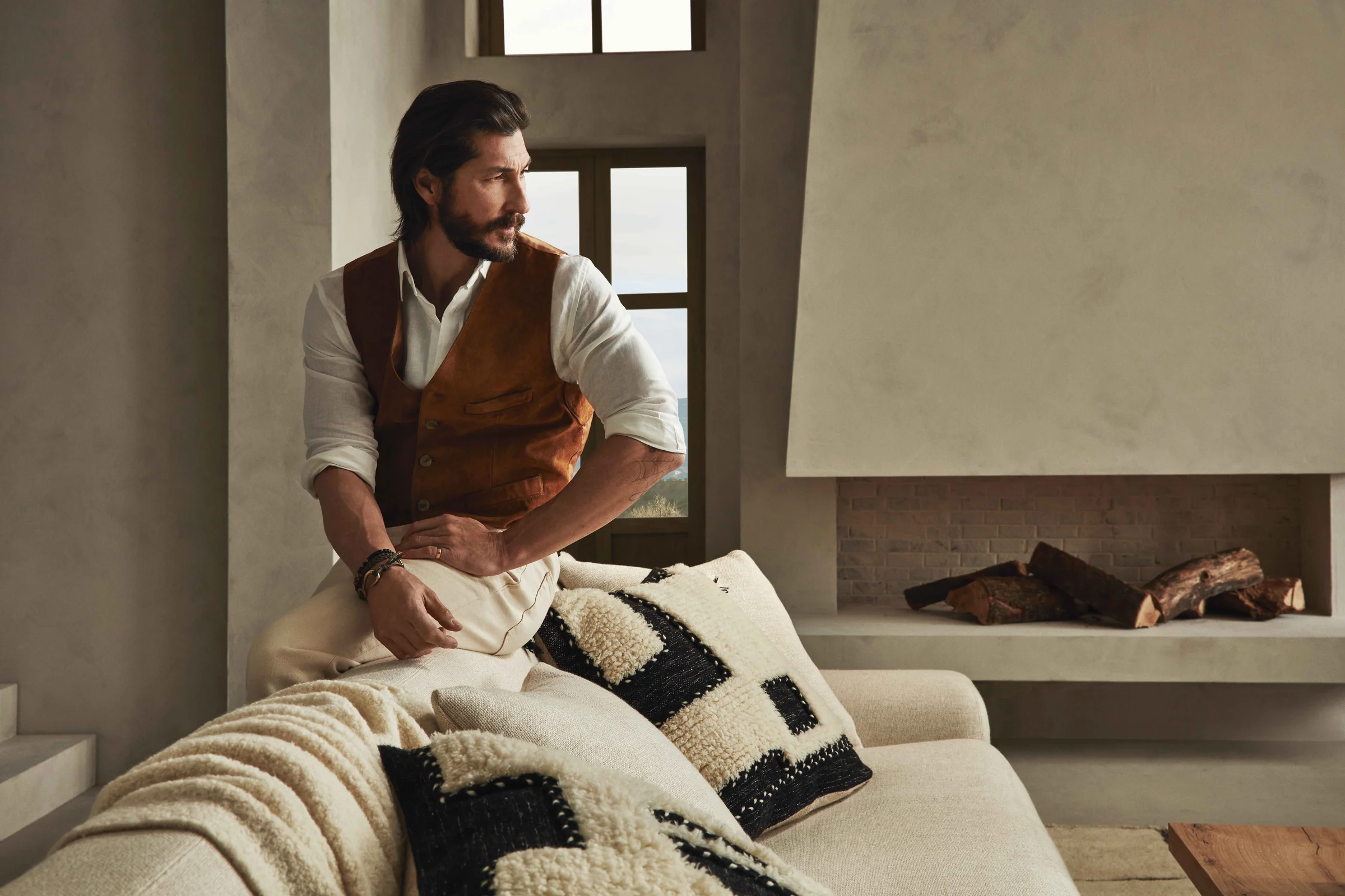 Bearded man in a tan suede vest and white shirt sits on a cream sofa with woven pillows near a fireplace, for Banana Republic.