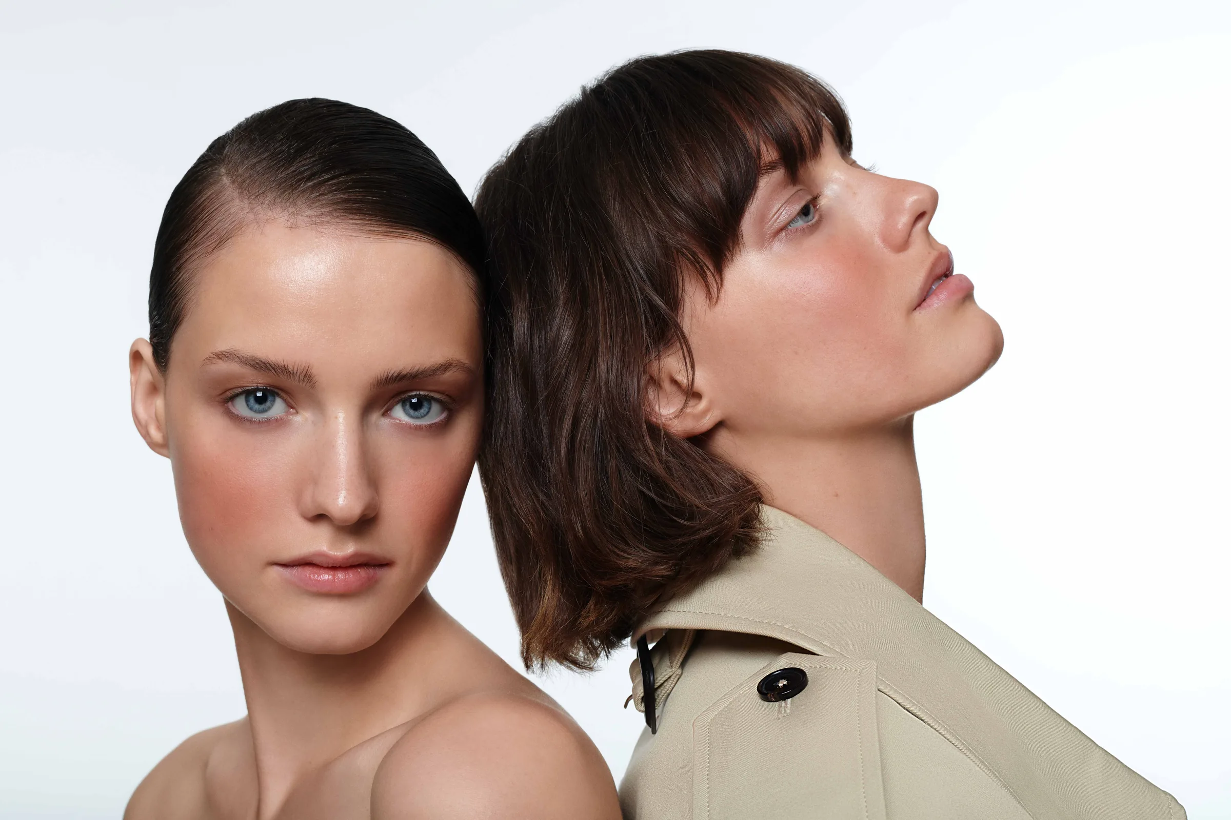 Two models posed back to back on white, one with slicked dark hair facing forward, one in a beige trench coat in profile.
