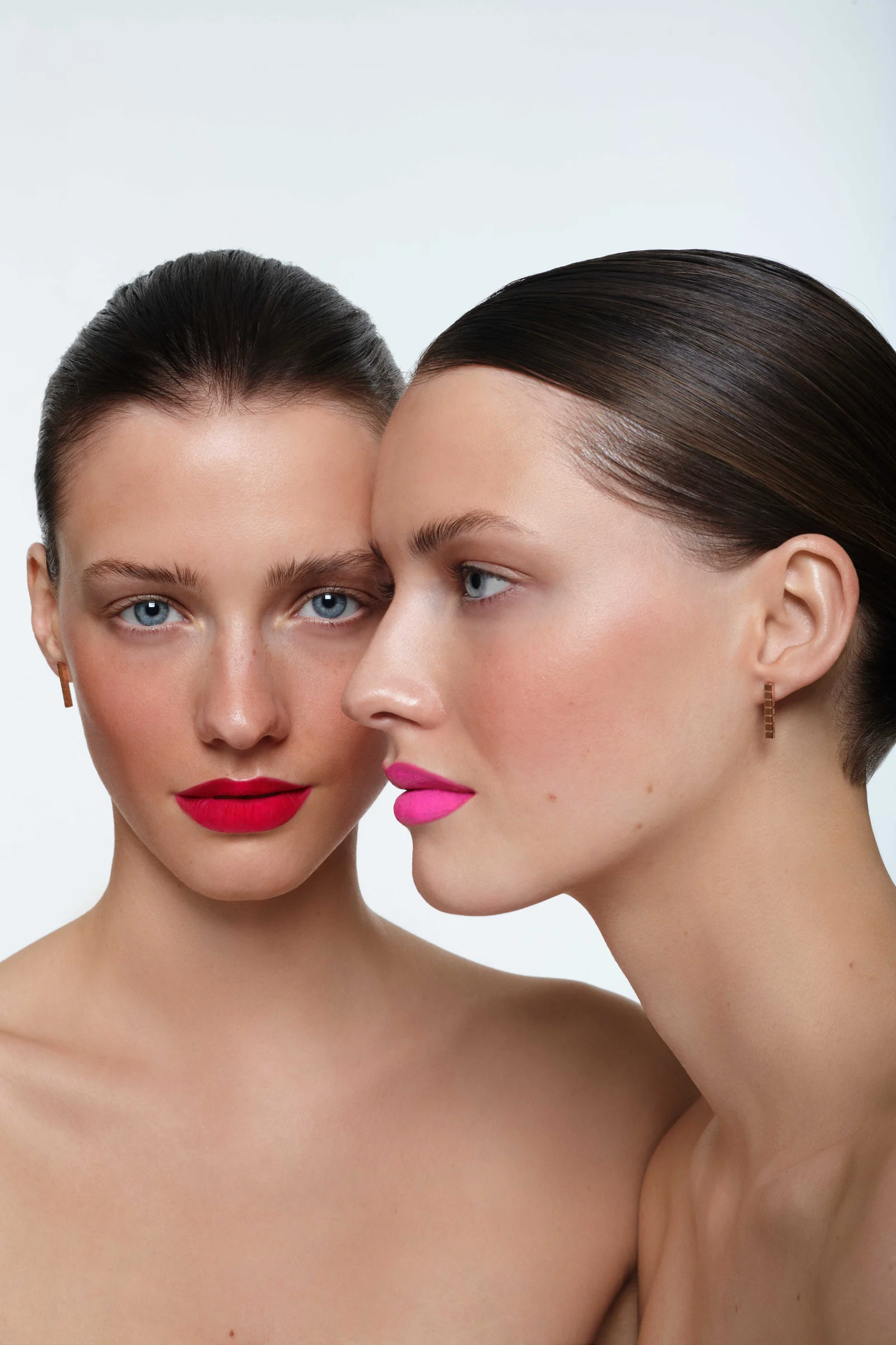 Two models with slicked-back hair, one in red matte lipstick facing forward, one in pink lipstick in profile.