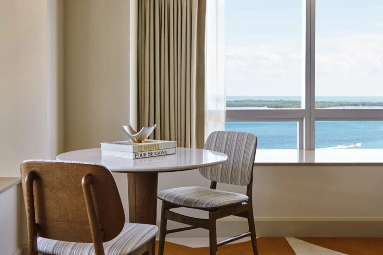 Hotel room corner with round white table, striped chairs and a window overlooking blue bay water, for Four Seasons.