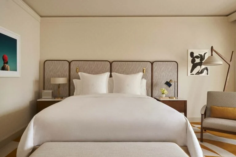 Hotel bedroom with white bedding, caned panel headboard, brass lamps, grey armchair and framed art, for Four Seasons.