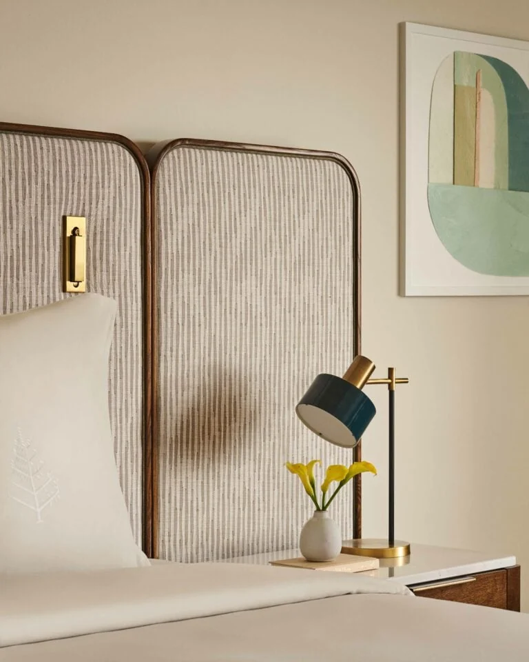 Hotel bed with striped upholstered headboard, brass sconce, teal lamp and yellow calla lilies on nightstand, for Four Seasons.