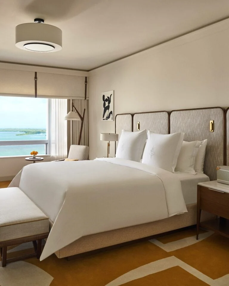 Hotel suite bedroom with white king bed, woven headboard, ochre rug and window overlooking the bay, for Four Seasons.