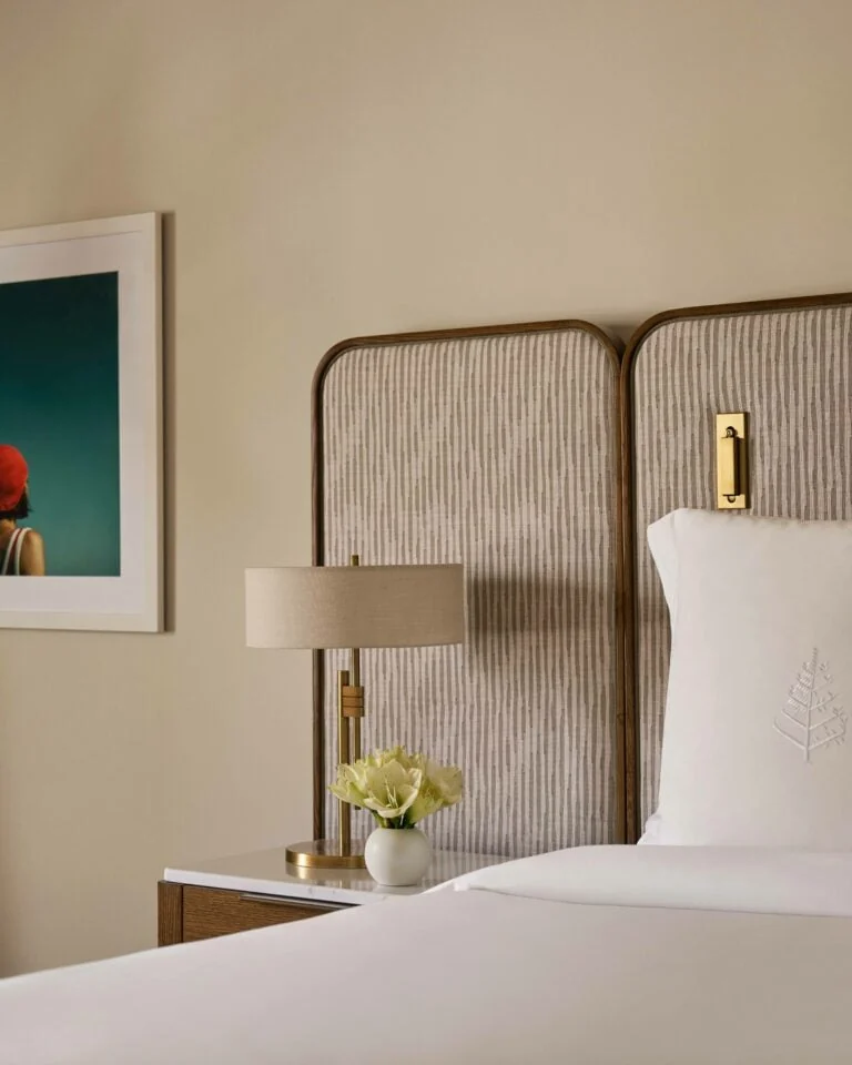 Hotel bed with striped upholstered headboard, embroidered pillow, brass lamp and white flowers on nightstand, for Four Seasons.