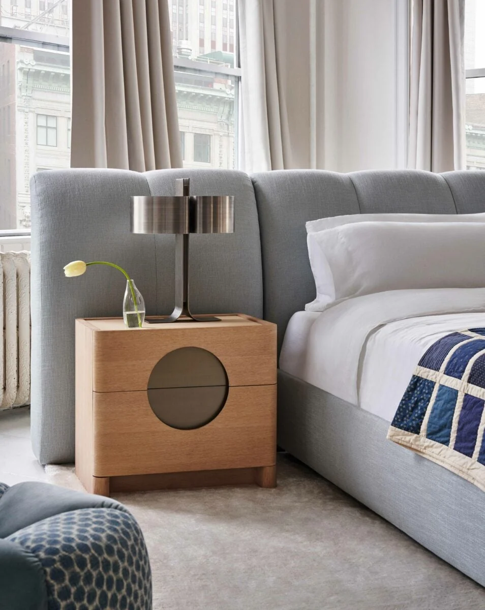 Bedroom interior with oak nightstand, metal table lamp and grey upholstered bed beside tall curtained windows.