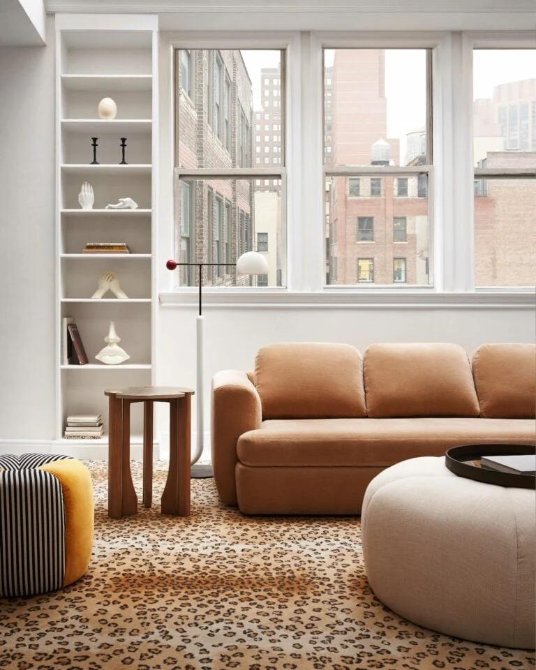 Living room with camel velvet sofa, leopard-print carpet, round ottoman and built-in shelves before tall city windows.