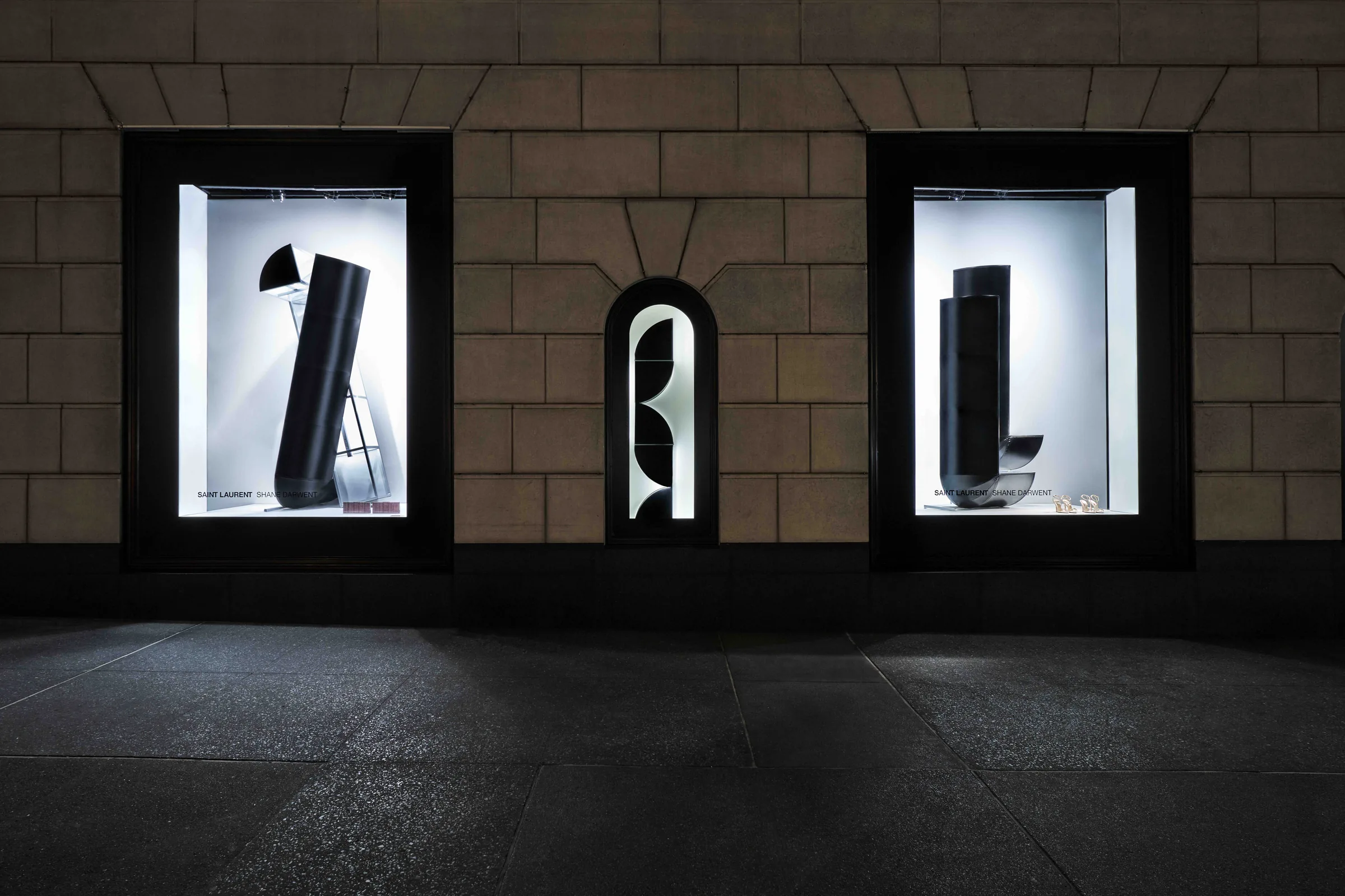 Storefront windows at night with black sculptural forms lit against white backdrops in a stone facade, for Saint Laurent.