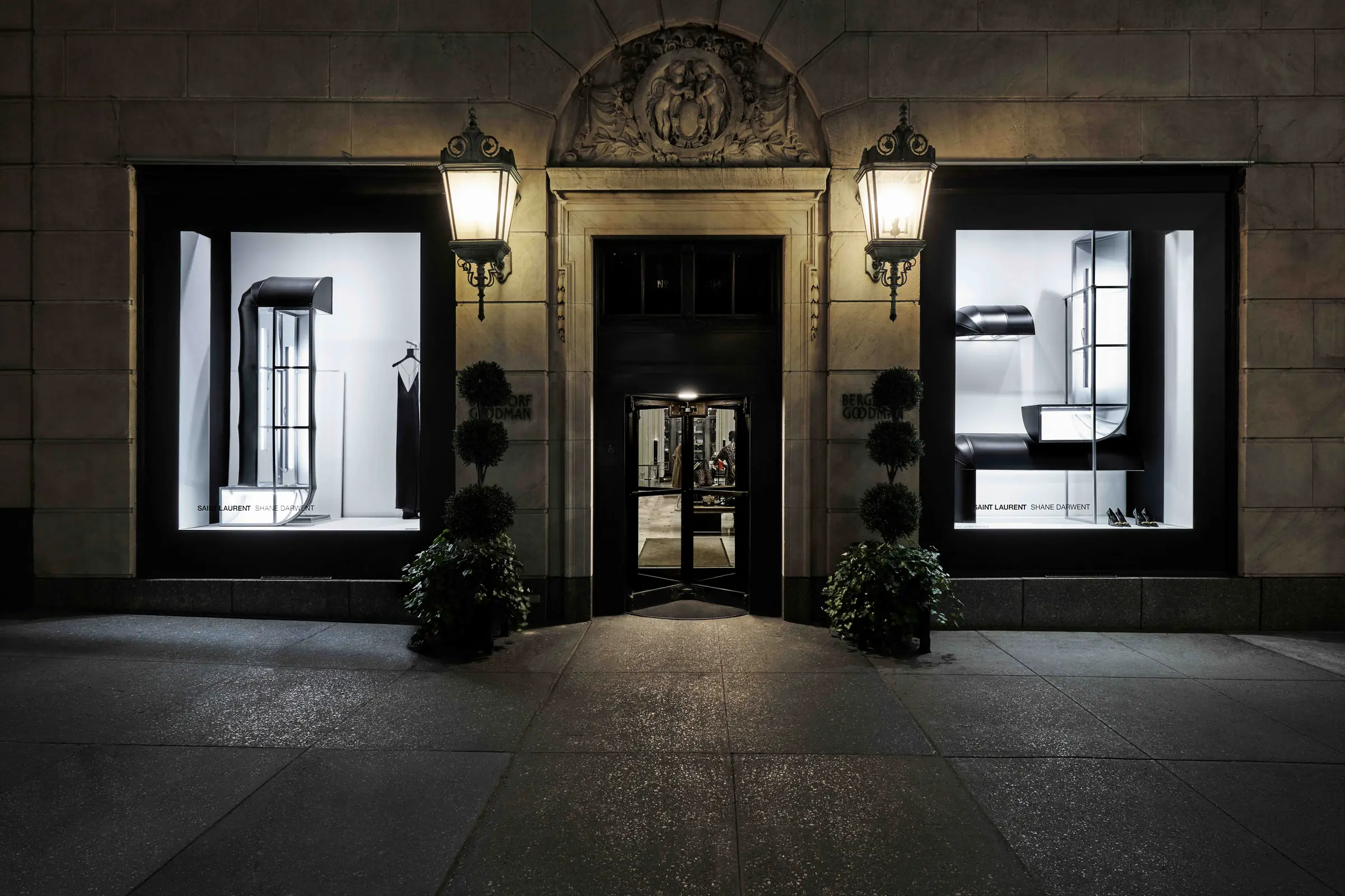 Stone storefront at night with lit window displays, lanterns and topiaries flanking the entrance, for Saint Laurent.