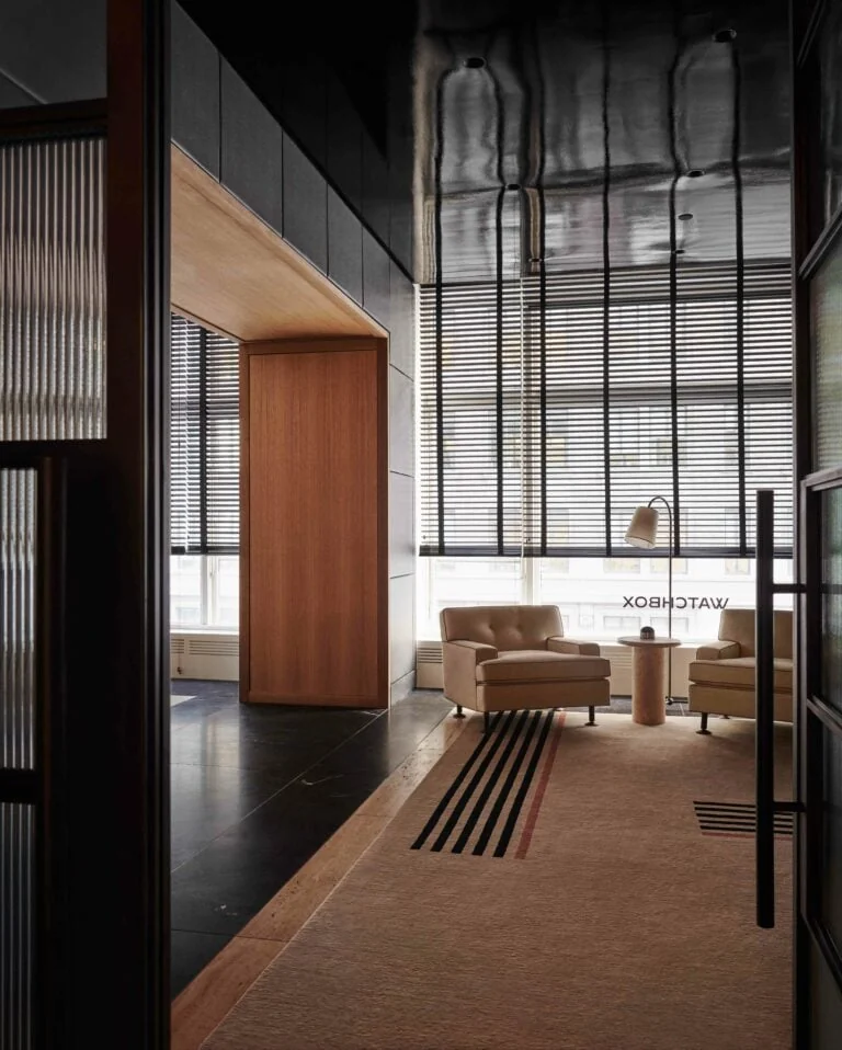 Interior lounge with beige tufted armchairs, striped rug, wood-clad column and slatted window blinds, for WatchBox.