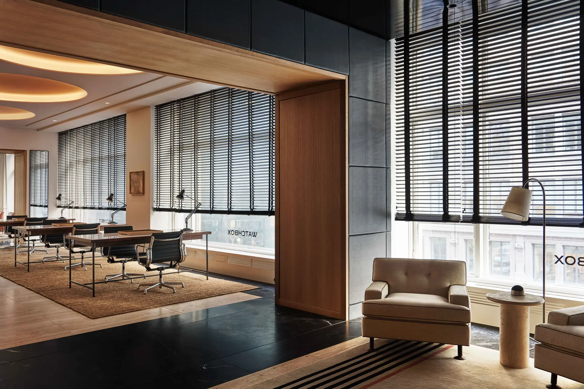 Interior with beige armchairs, wood-framed opening and desks with black chairs before tall window blinds, for WatchBox.