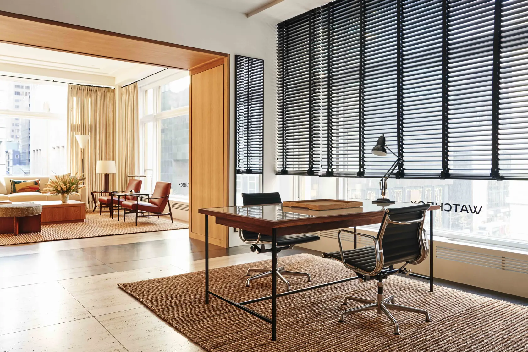 Office interior with glass-top desk, black leather chairs, jute rug and lounge beyond tall bladed windows, for WatchBox.