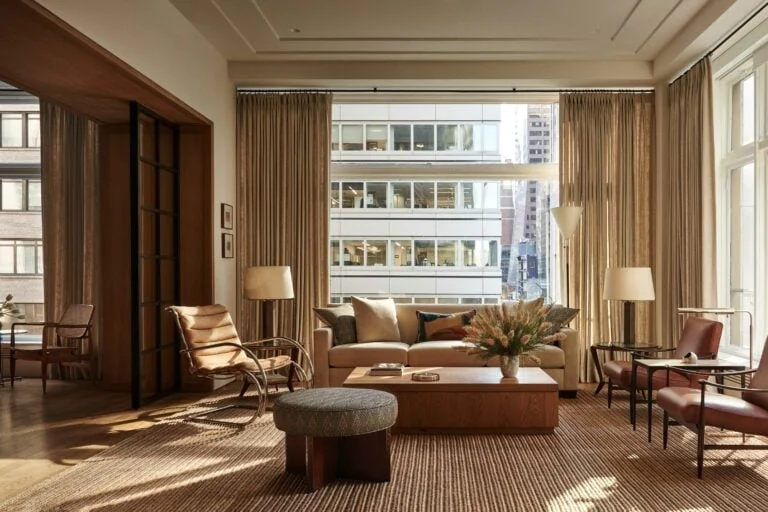 Sunlit interior lounge with beige sofa, leather lounge chairs and tall curtains overlooking city offices, for WatchBox.