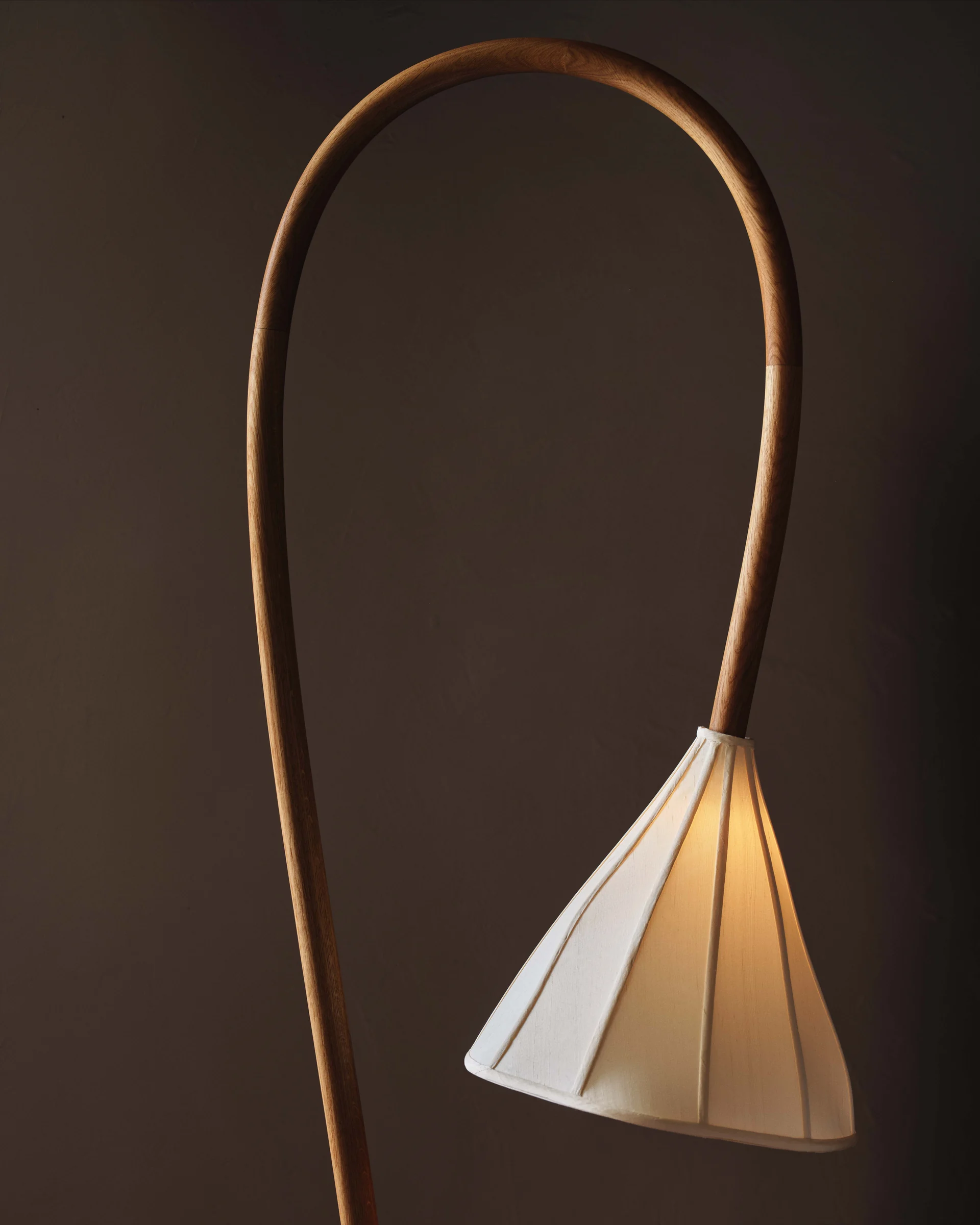 Arched bentwood floor lamp with a glowing pleated linen shade against a dark brown wall, for Anna Karlin.