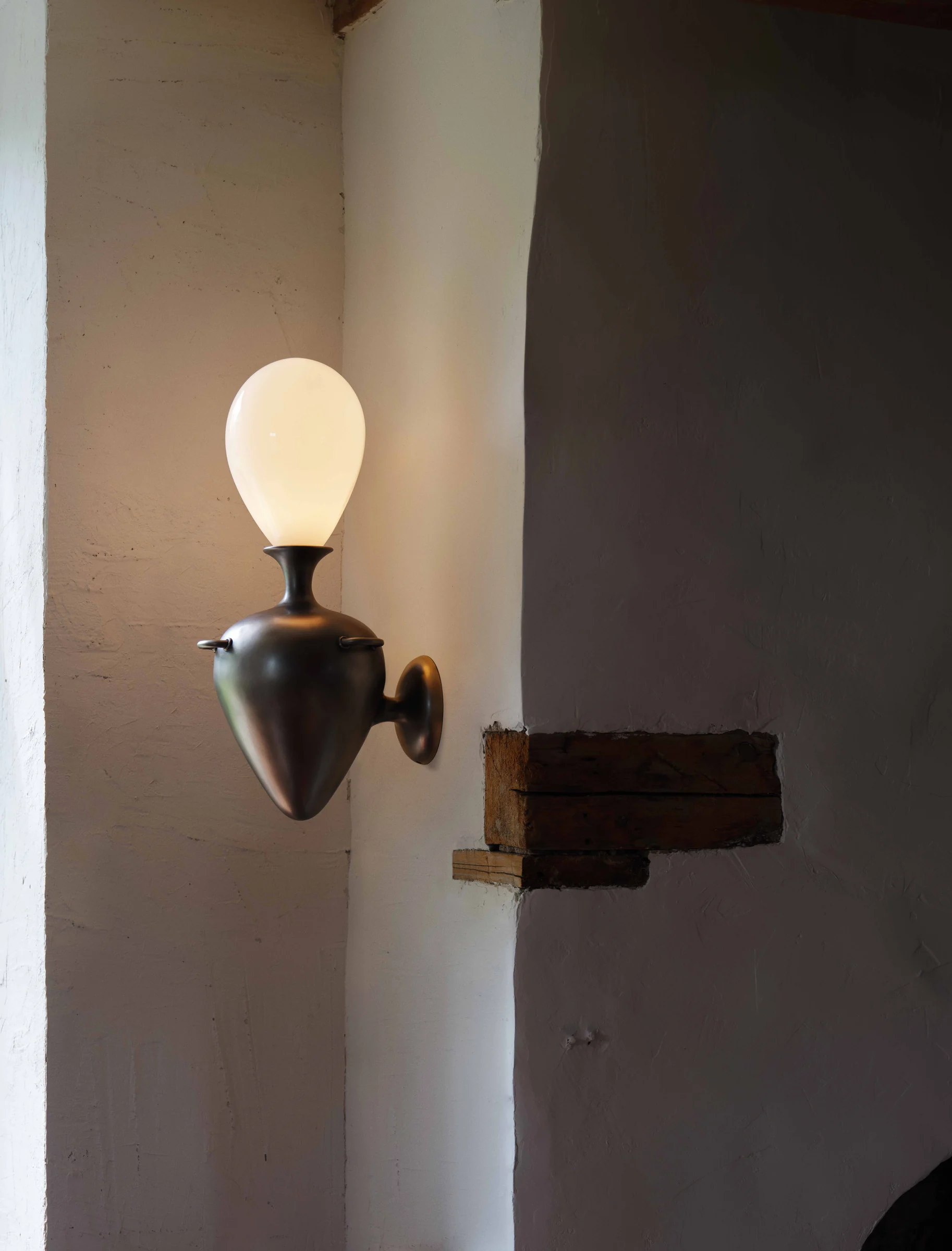 Bronze wall sconce with a glowing opal glass bulb on a whitewashed plaster wall beside a timber beam, for Anna Karlin.