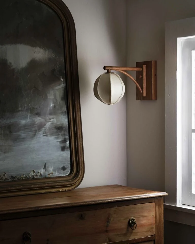 Wooden wall sconce with a round cream shade beside an antique mirror and wooden dresser in window light, for Anna Karlin.
