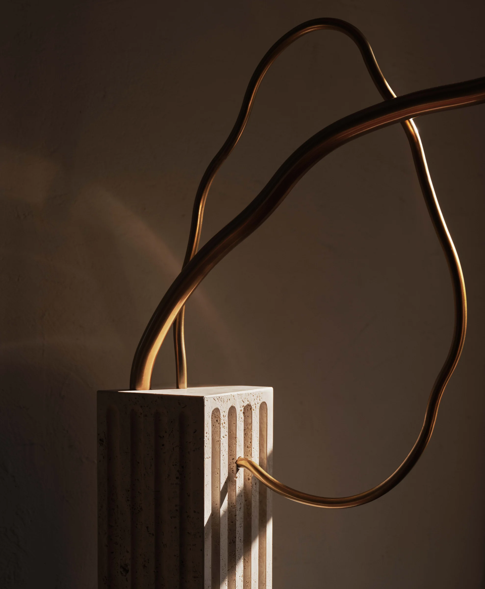 Curving brass tube sculpture rising from a fluted travertine base in warm raking light, for Anna Karlin.