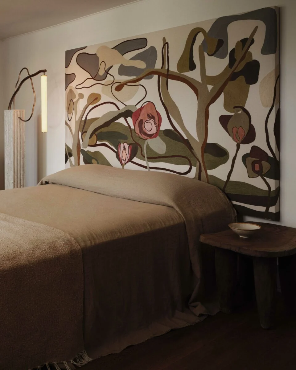 Bedroom with a taupe linen bedspread, abstract floral wall tapestry, sculptural lamp and wooden side table, for Anna Karlin.