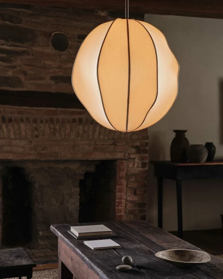 Glowing silk pendant lamp above a rustic wooden table with books and stones before a brick fireplace, for Anna Karlin.