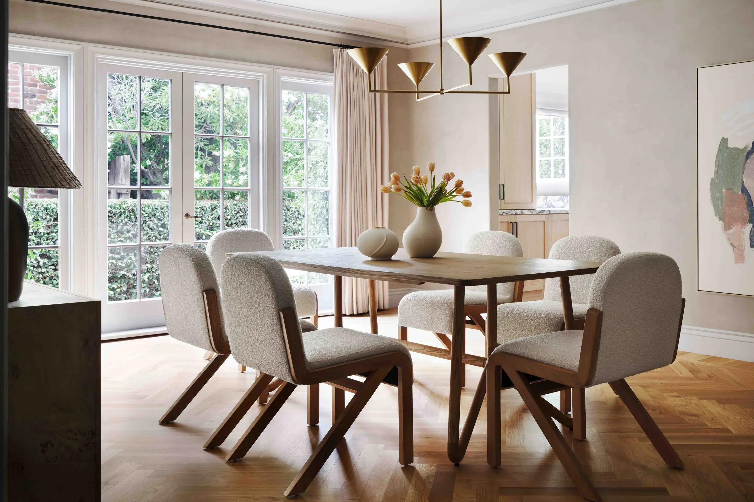 Dining room with oak table, cream bouclé chairs, brass pendant and tulips by French doors, for Anthropologie.